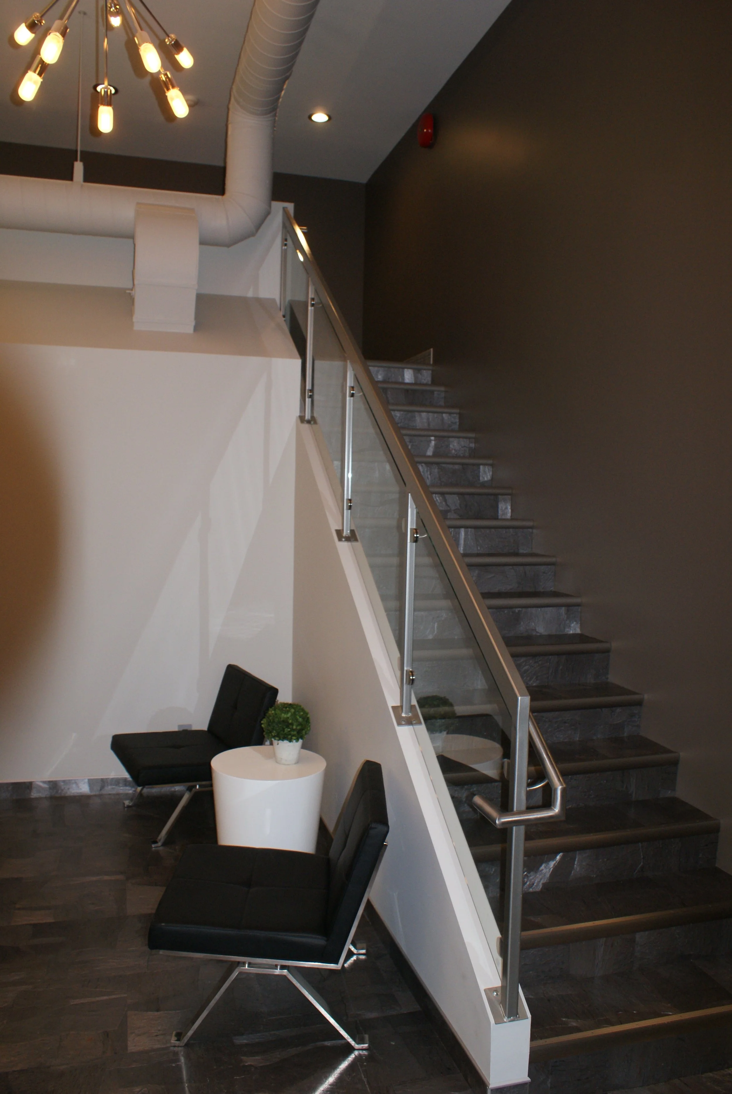 Interior of a modern building with a staircase featuring dark stone steps and glass railing, black leather chairs, a white round side table with a green potted plant, dark wooden flooring, and a ceiling light fixture with multiple exposed bulbs.