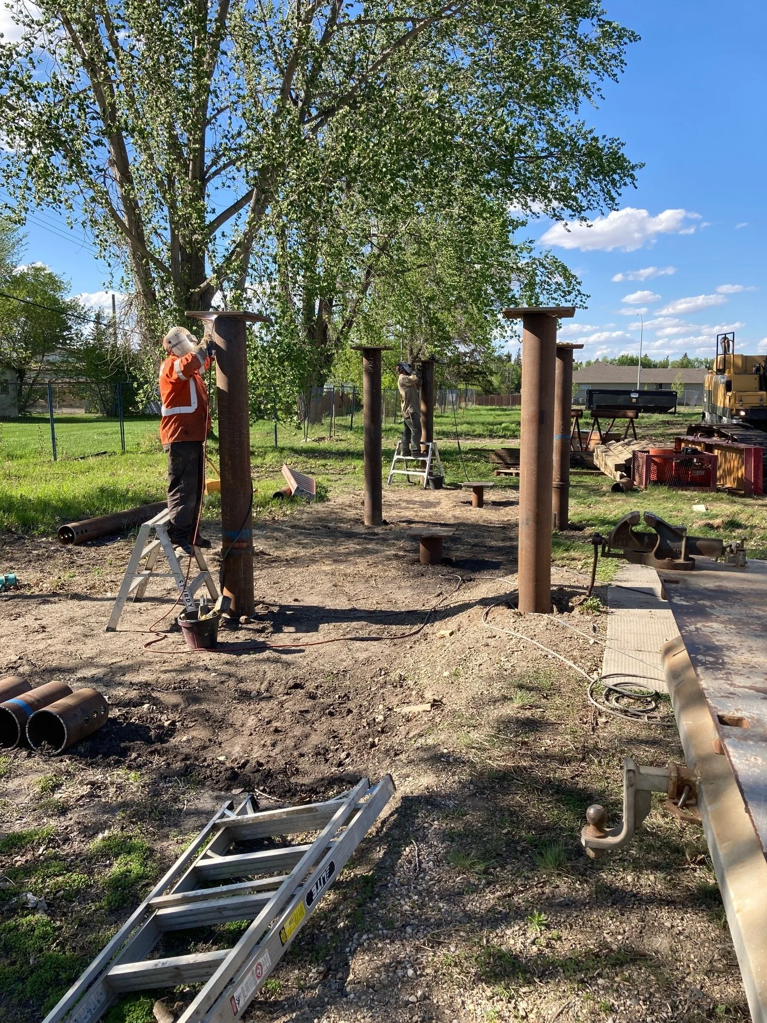 Two workers installing or repairing metal poles outdoors on a sunny day, with a large tree and green grass in the background.