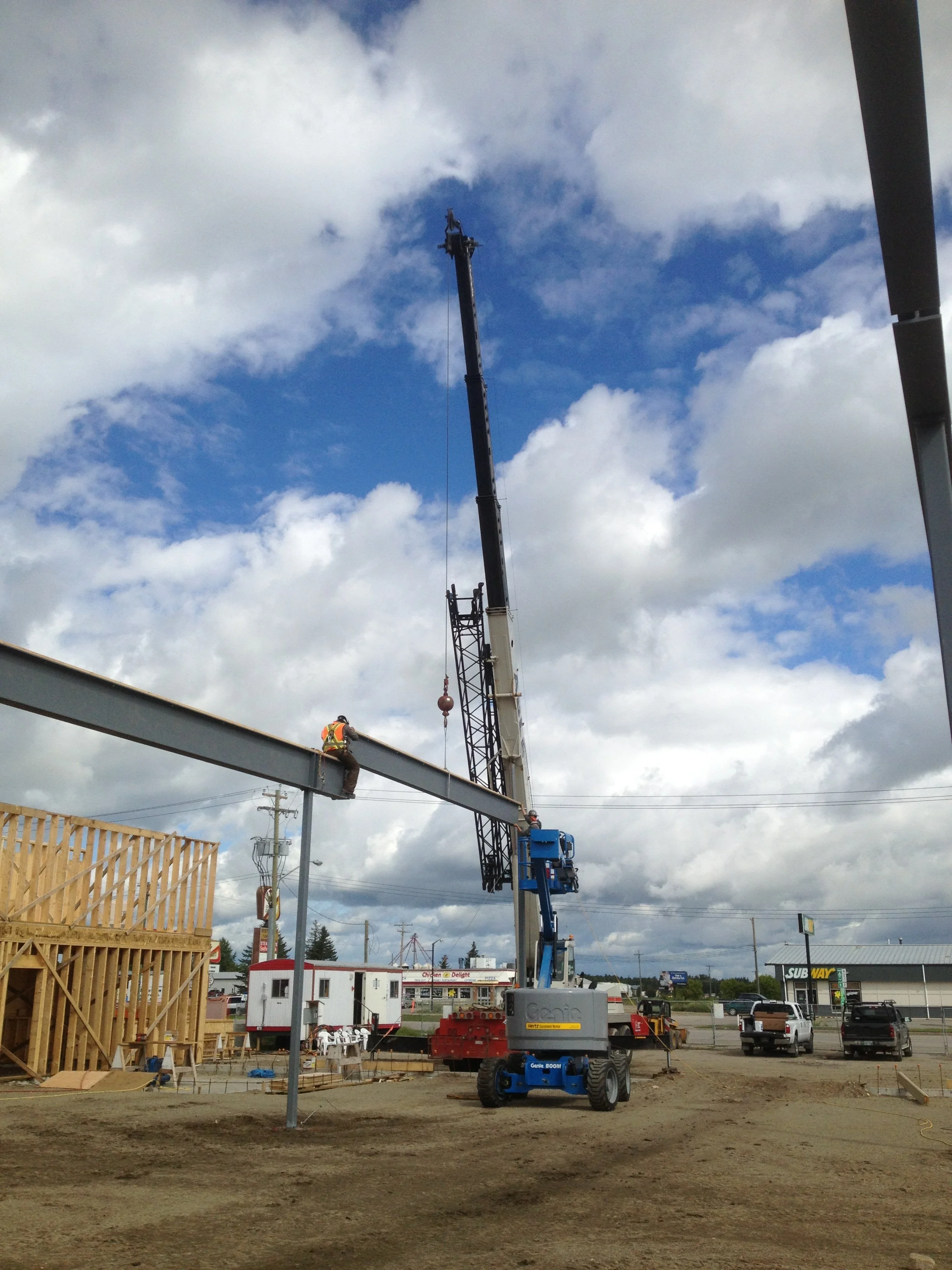 Construction workers on a steel beam with a crane lifting materials at a building site, with parking lot and Subway restaurant in the background.