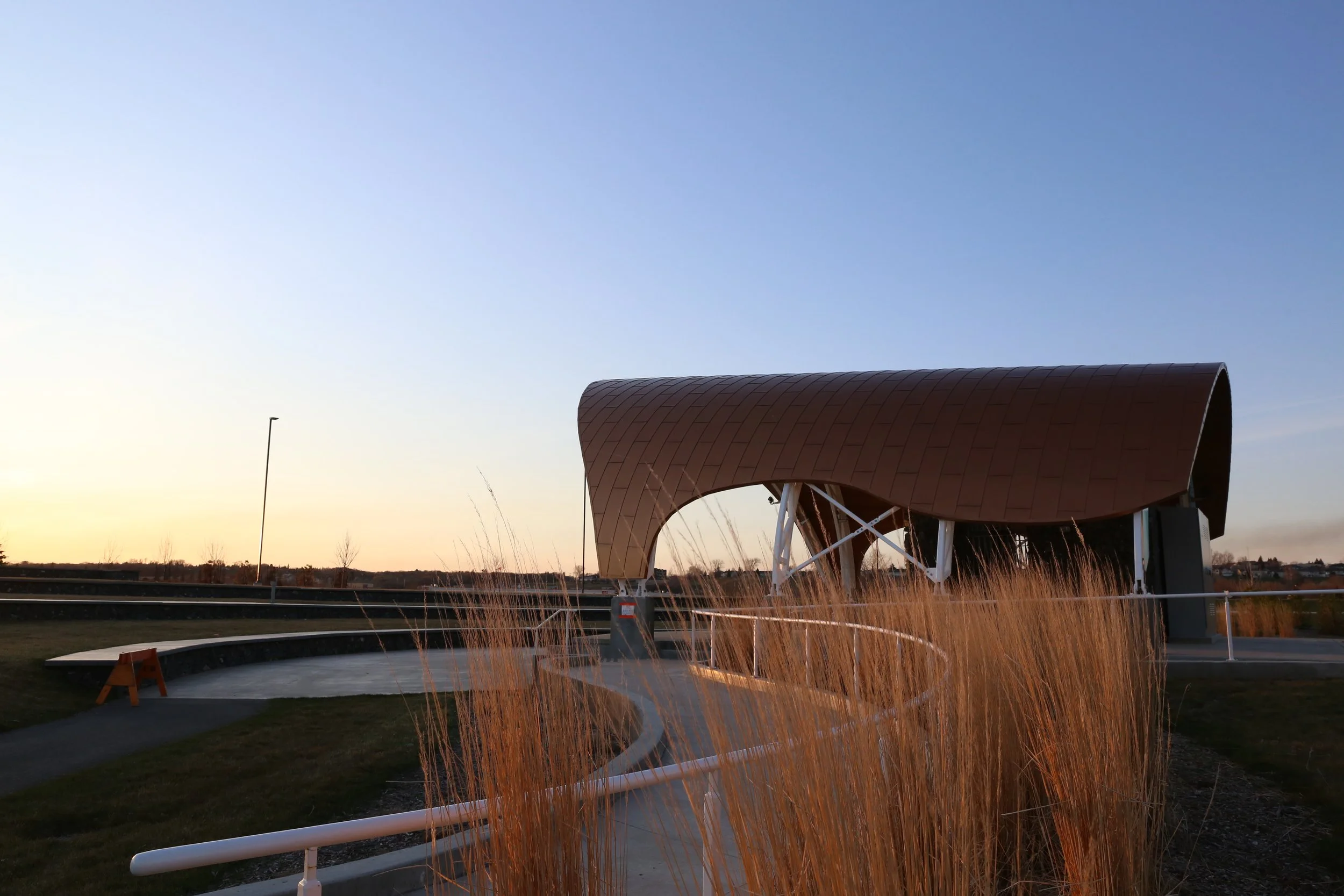 Outdoor park with modern structure featuring a curved brown roof, white support beams, walking path, dry grasses, and a sunset sky.