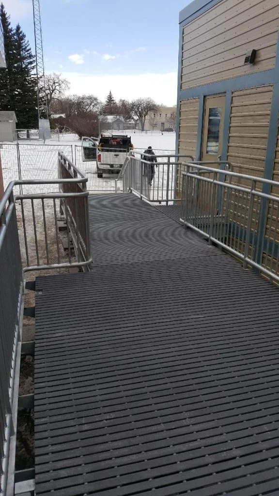 Metal ramp with handrails outside a building, with snowy ground and parked vehicles in the background.