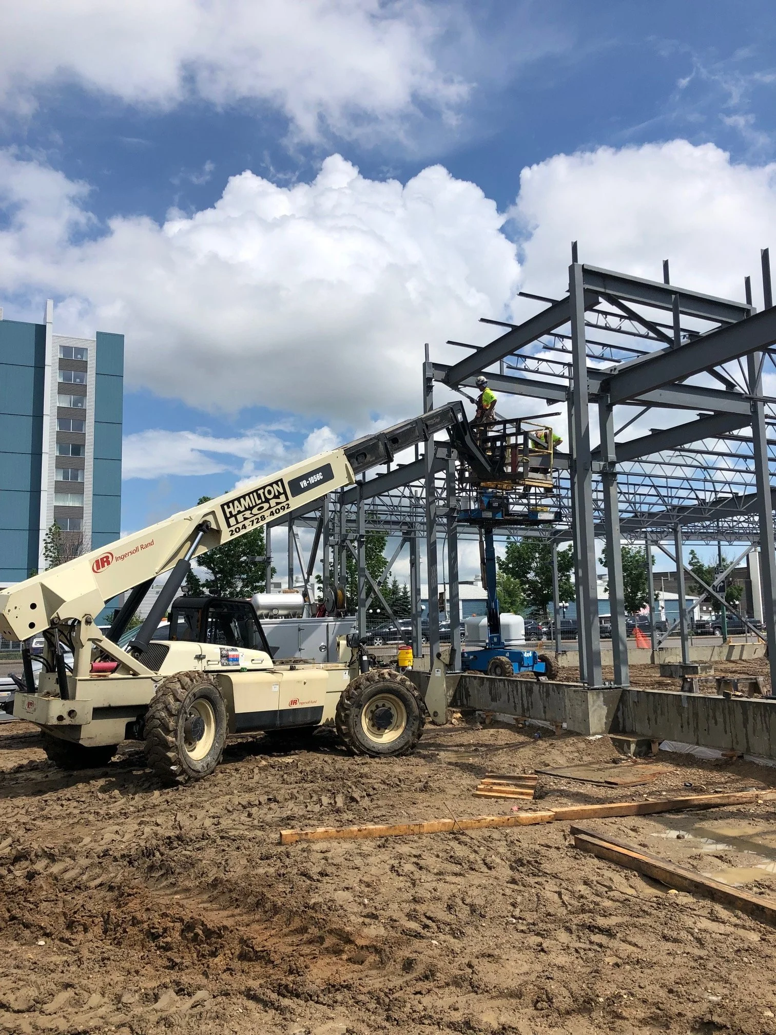 Construction workers are building a steel framework structure using a crane on a construction site with a dirt ground and a partly cloudy sky.