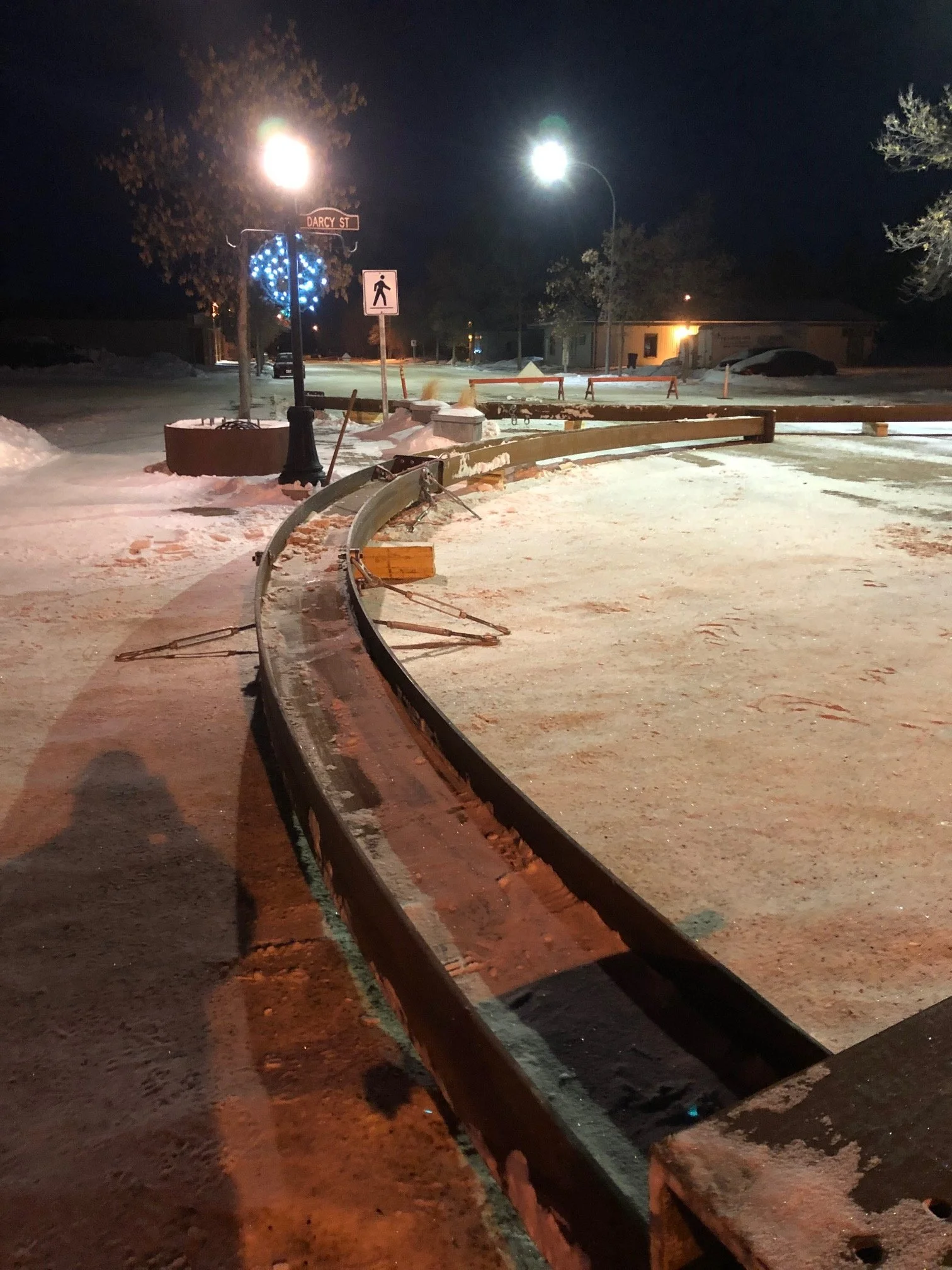 A damaged metal guardrail on a snow-covered street at night, illuminated by streetlights and a decorative blue lighted heart-shaped installation, with a pedestrian crossing sign and trees in the background.