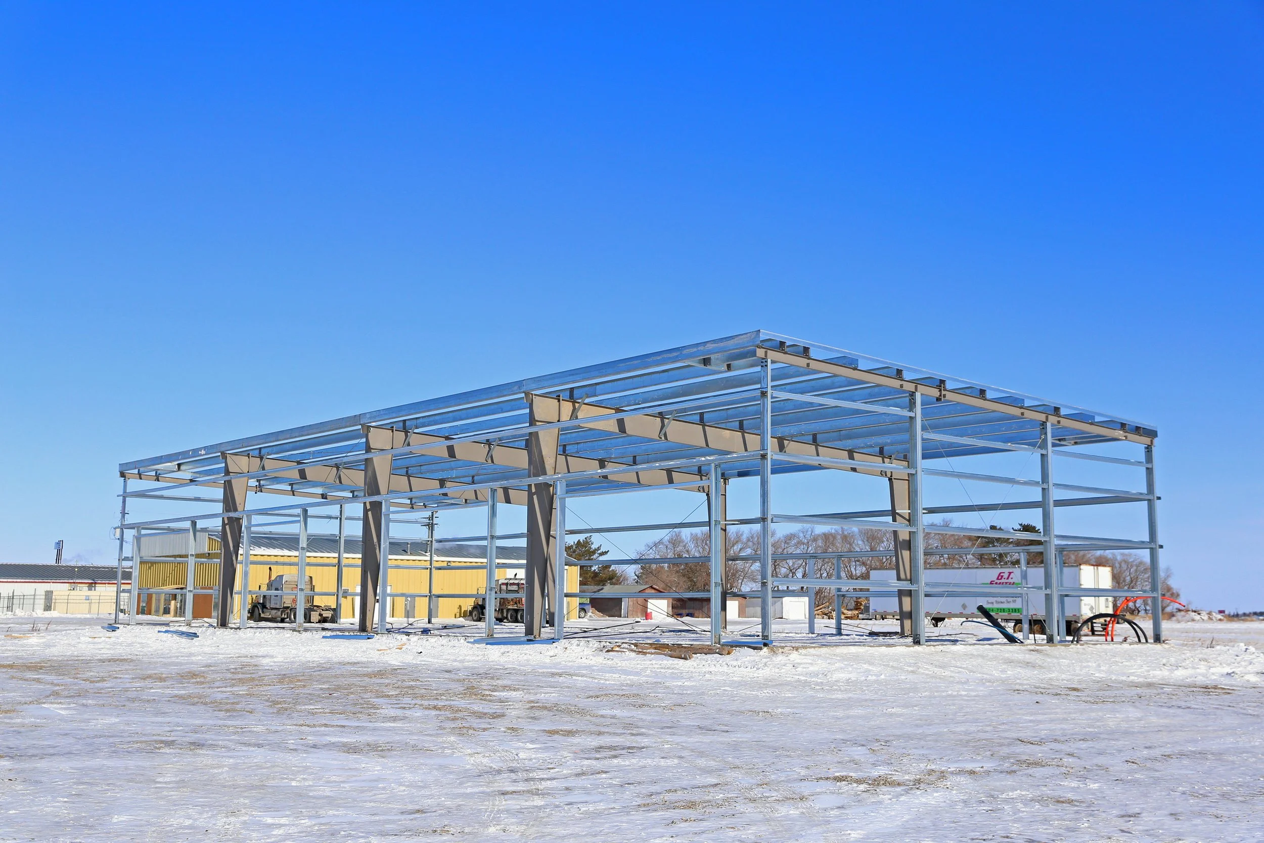 Steel frame structure under construction on snowy ground with a clear blue sky.
