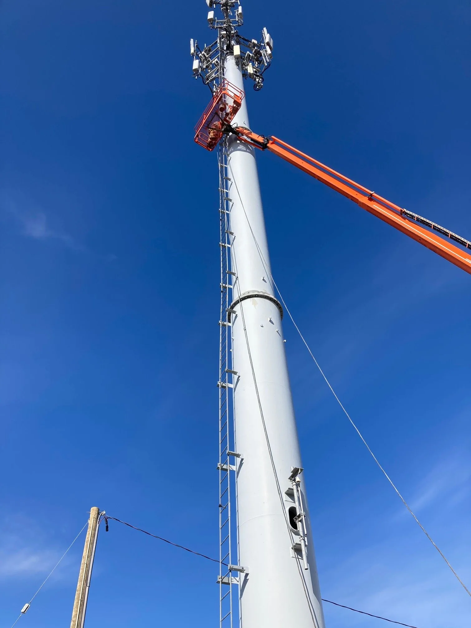 A telecommunications tower being serviced by a cherry picker lift with an orange extendable arm, against a clear blue sky.