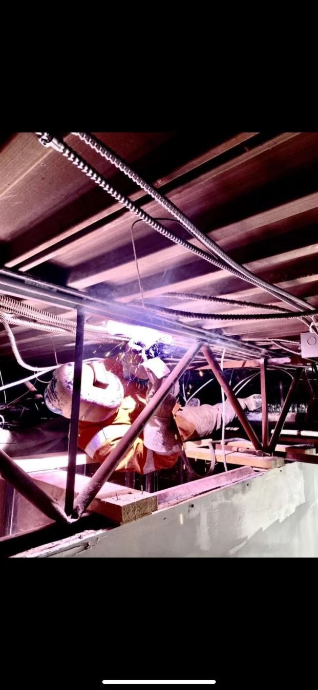 Construction worker welding metal framework beneath a wooden structure, with sparks and bright light from welding.
