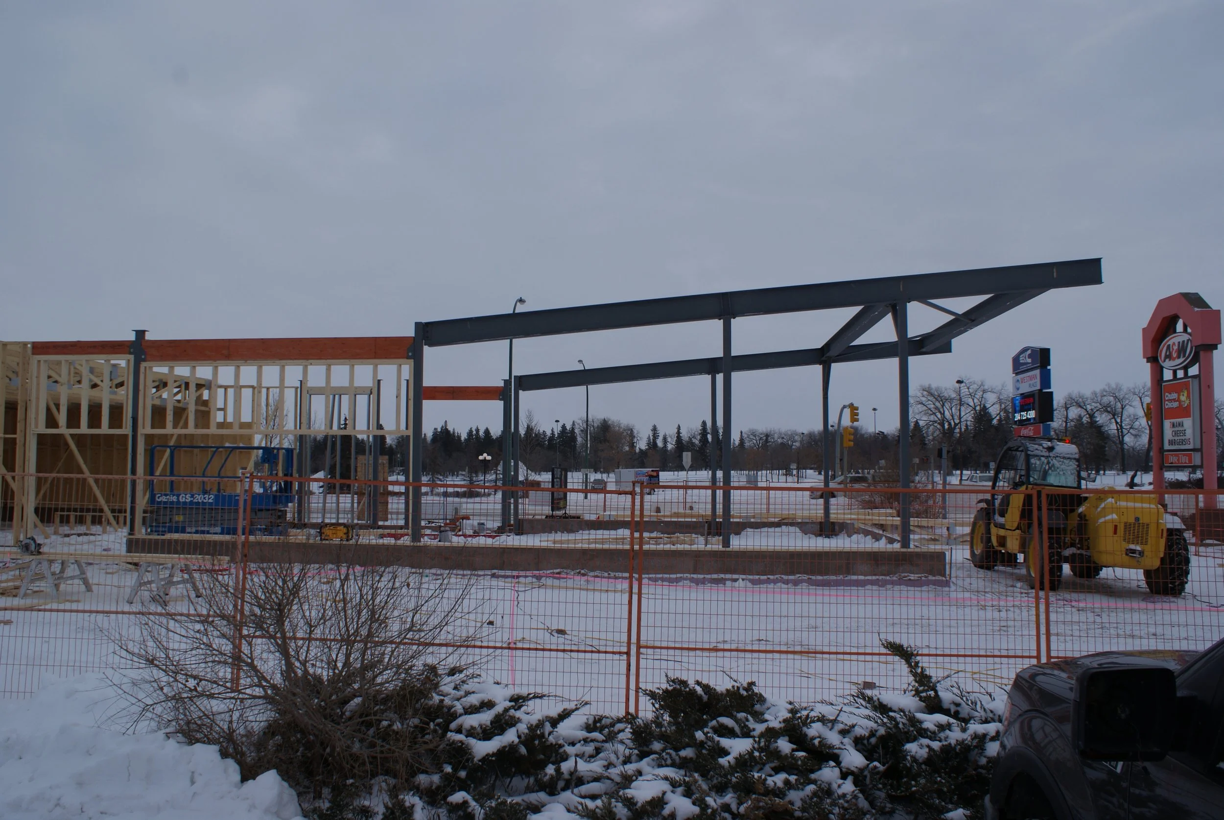 Construction site of a building with a steel frame and wooden walls, surrounded by snow, orange safety fencing, and construction equipment, near an A&W fast food restaurant.