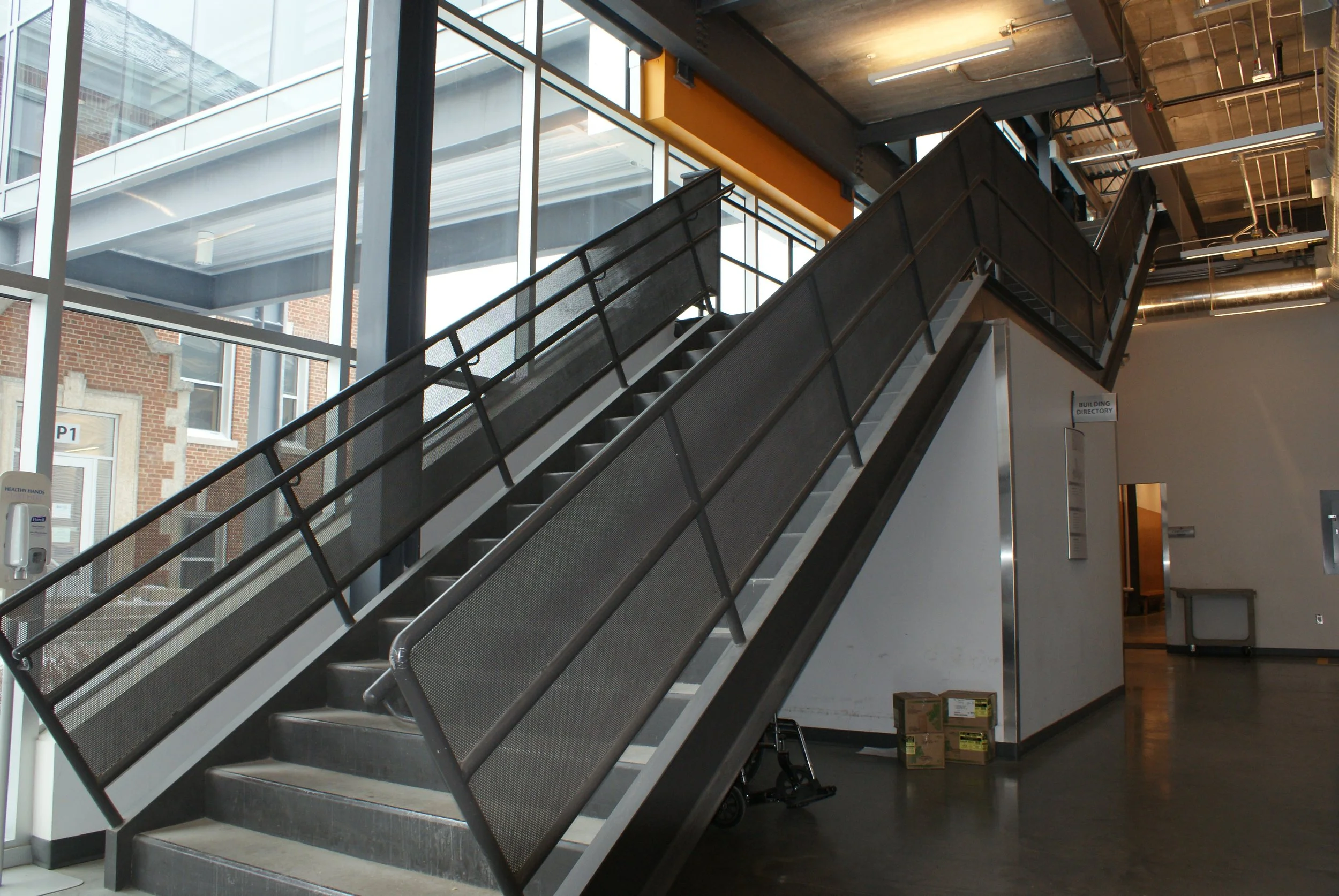 Indoor staircase with metal railings, leading to an upper level with large windows letting in natural light, in a modern industrial-style building.