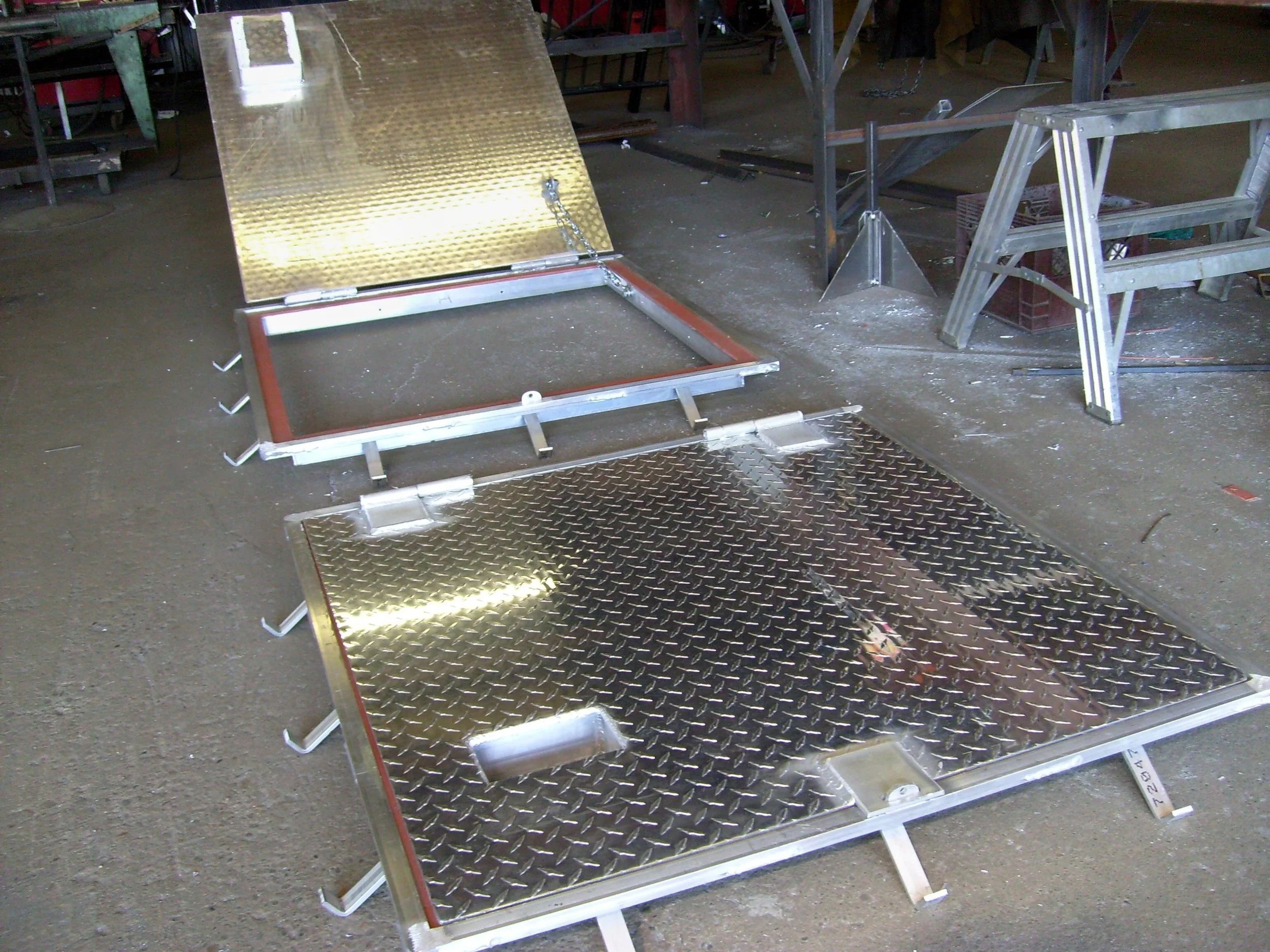 Metal hatch with diamond plate surface and a hinged lid, mounted on a frame with side brackets, placed on a concrete floor in a workshop.