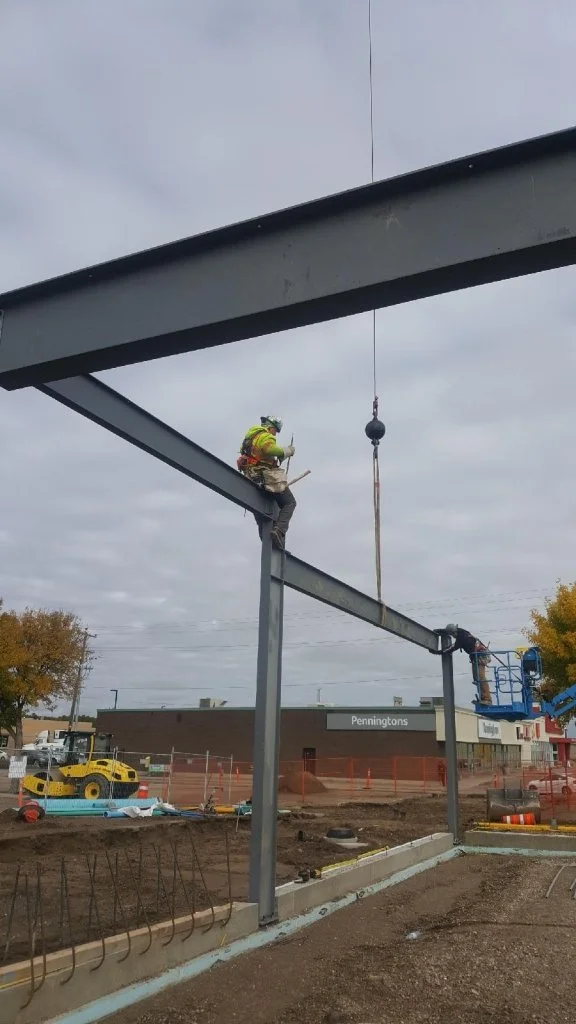 Construction worker in safety gear working on steel beams at a construction site with earth-moving equipment and a Pennington's store in the background.