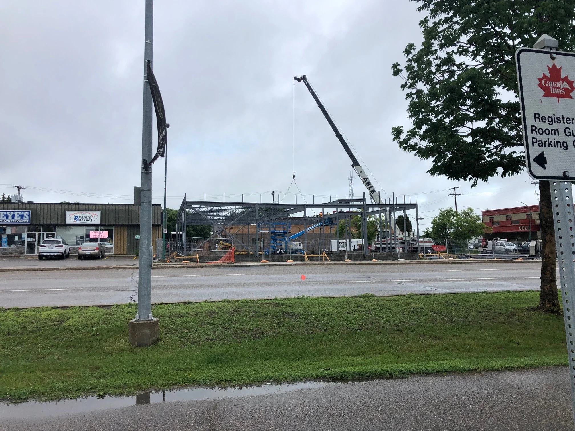 Construction site with a crane lifting framing for a building, parked cars, and a business storefront across the street.