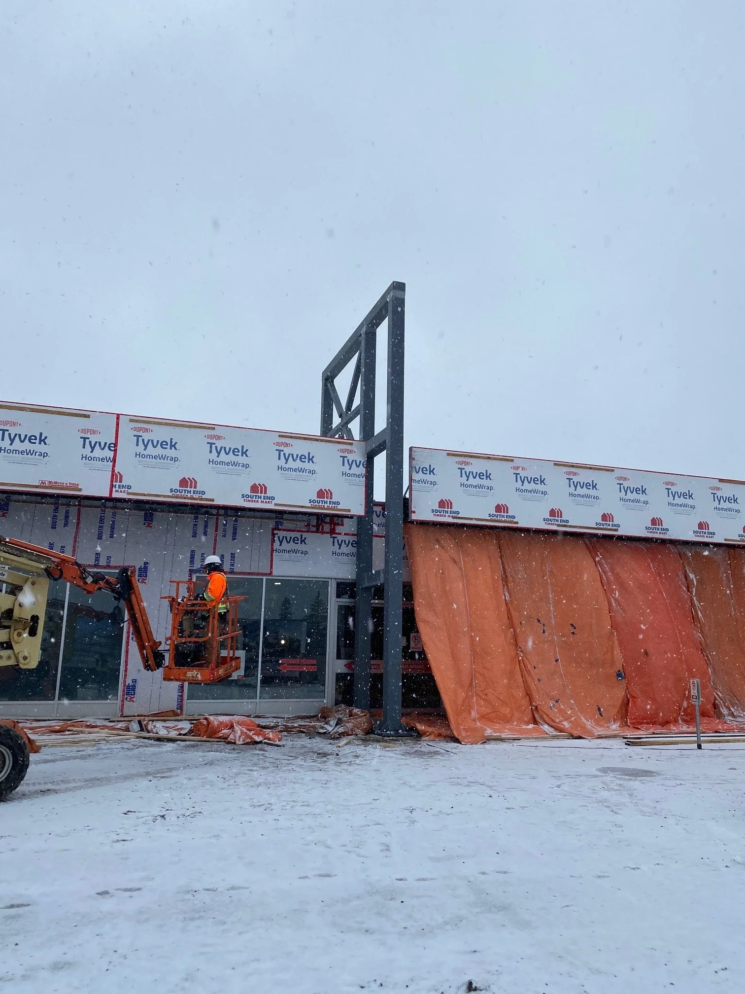 Construction worker in orange jacket and white helmet working on building exterior in snowy weather, with orange tarp covering part of the building wall.