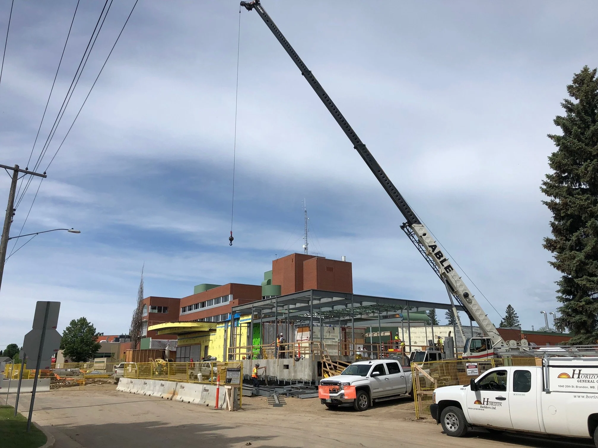 Construction site with workers and a crane lifting materials for building a structure, with trees and a multi-story brick building in the background.