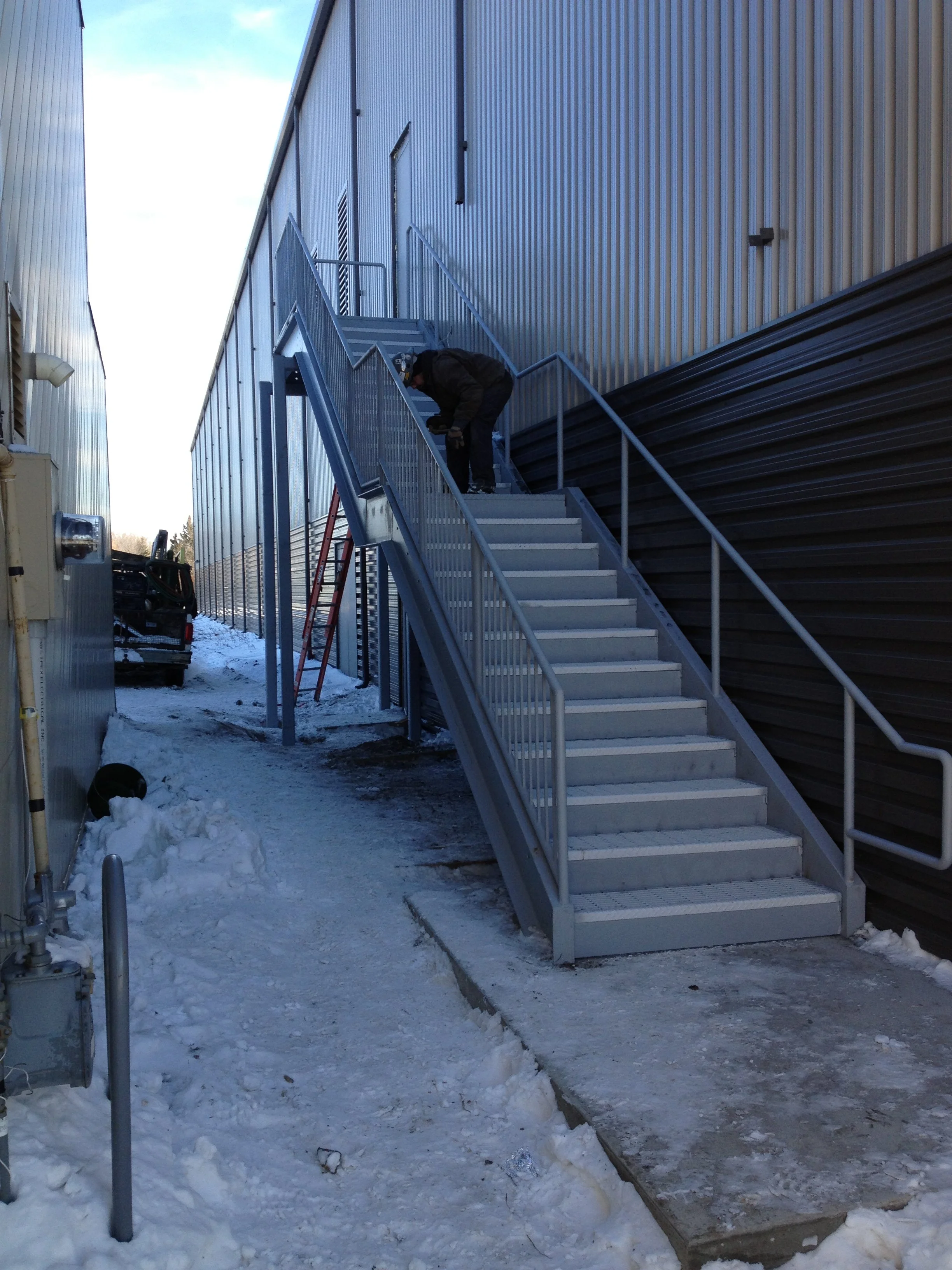 Man working on metal outdoor staircase attached to industrial building with snow on ground.