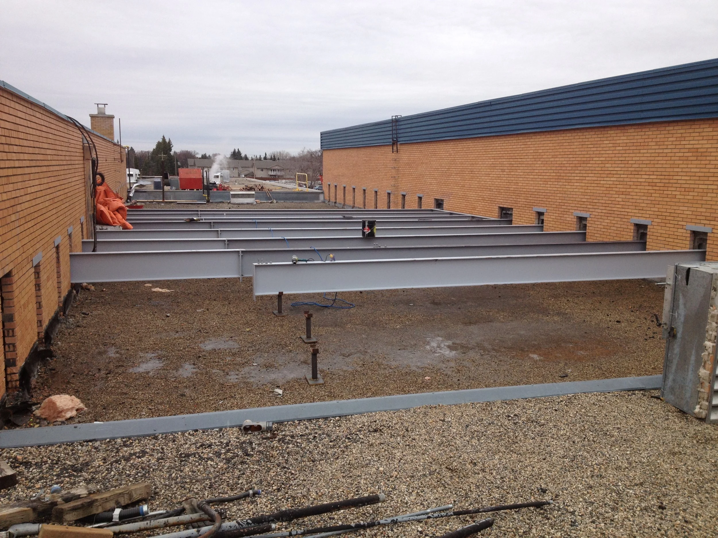 Construction site on a rooftop with metal beams and gravel, surrounded by brick walls, overcast sky.