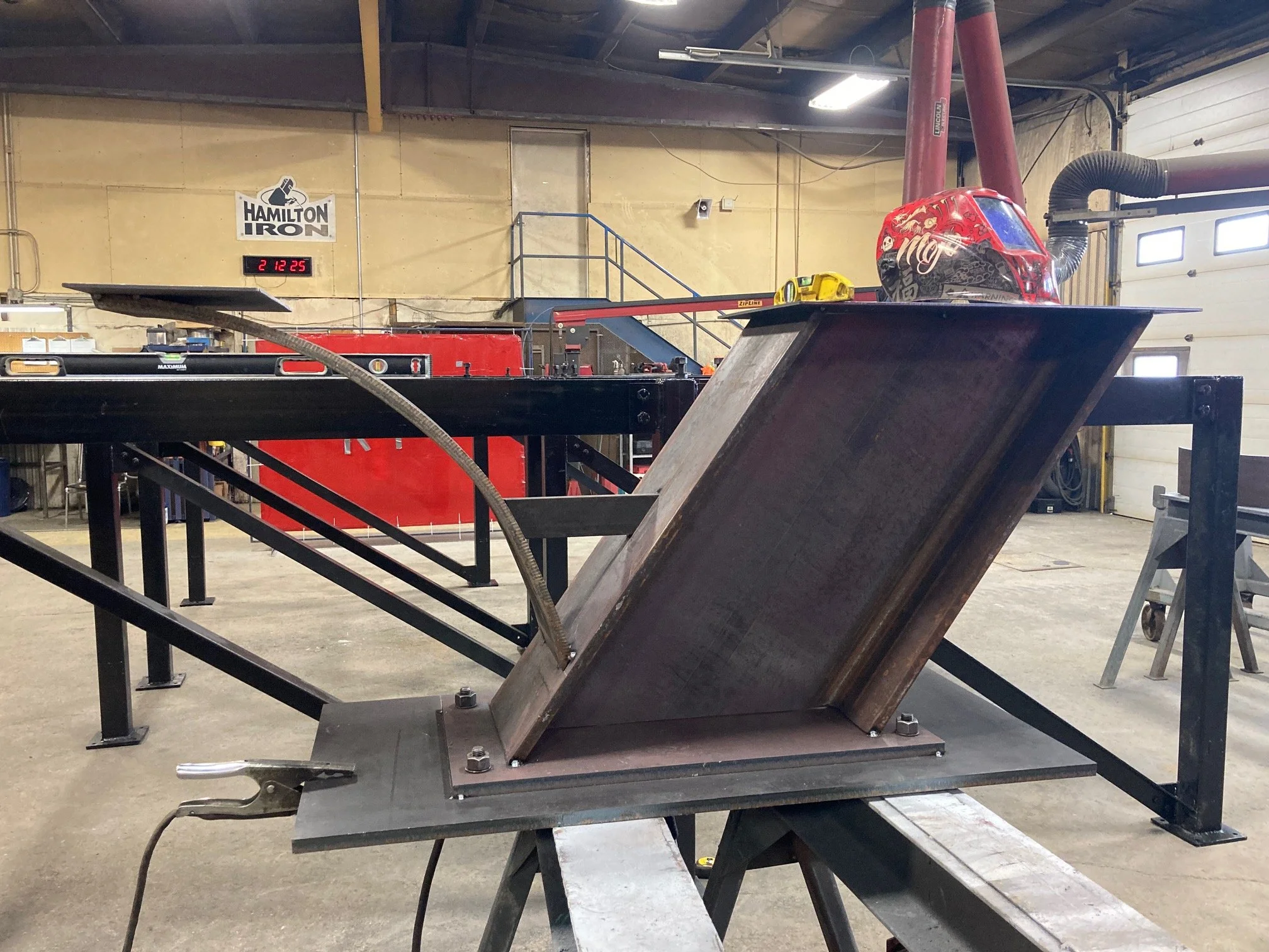 A welded metal structure in a workshop, with a helmet placed on top. The background shows a Hamilton Iron sign, red welding equipment, and a garage door.
