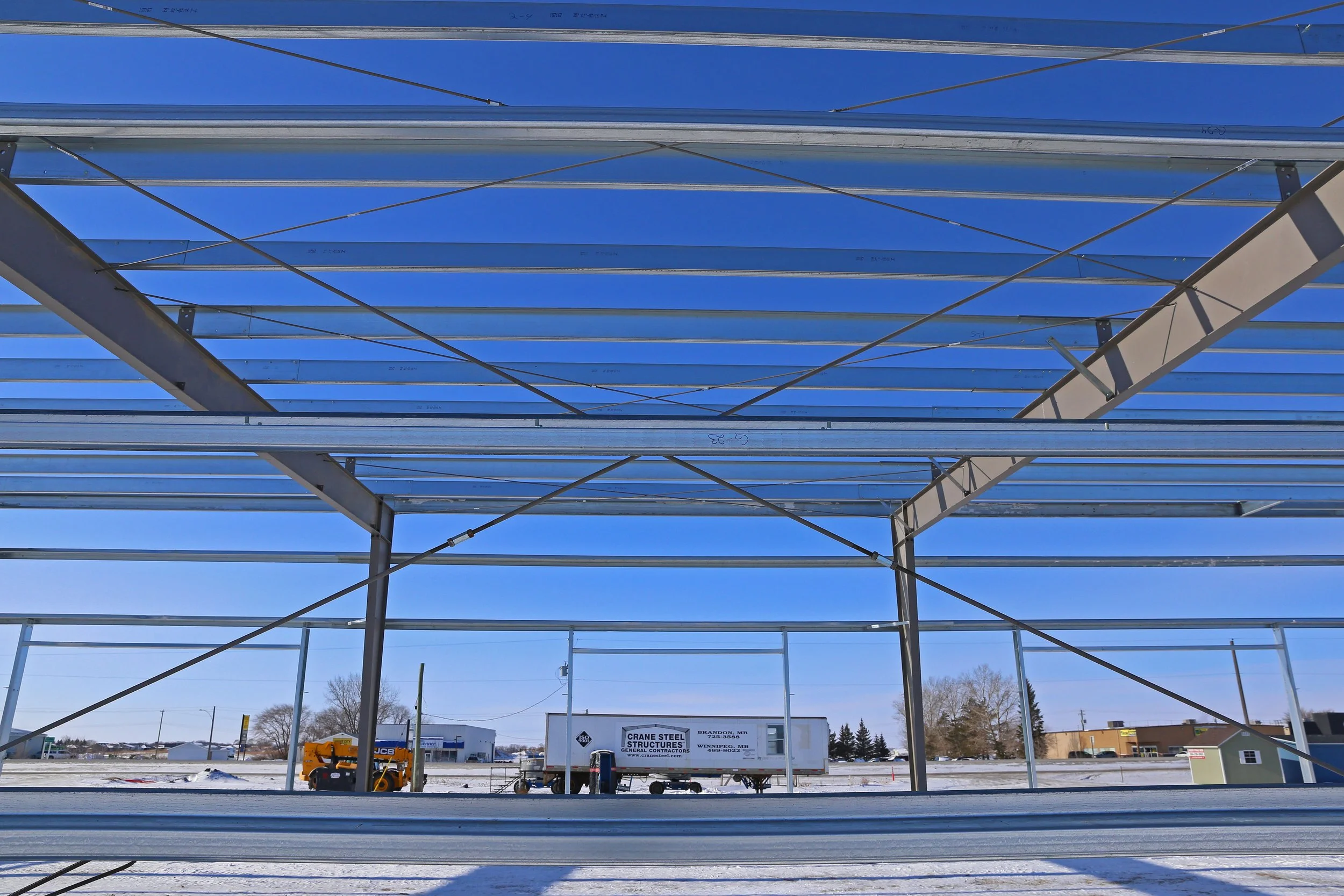 Metal framework under construction at an outdoor site with a clear blue sky, snow on the ground, and utility vehicles and buildings in the background.