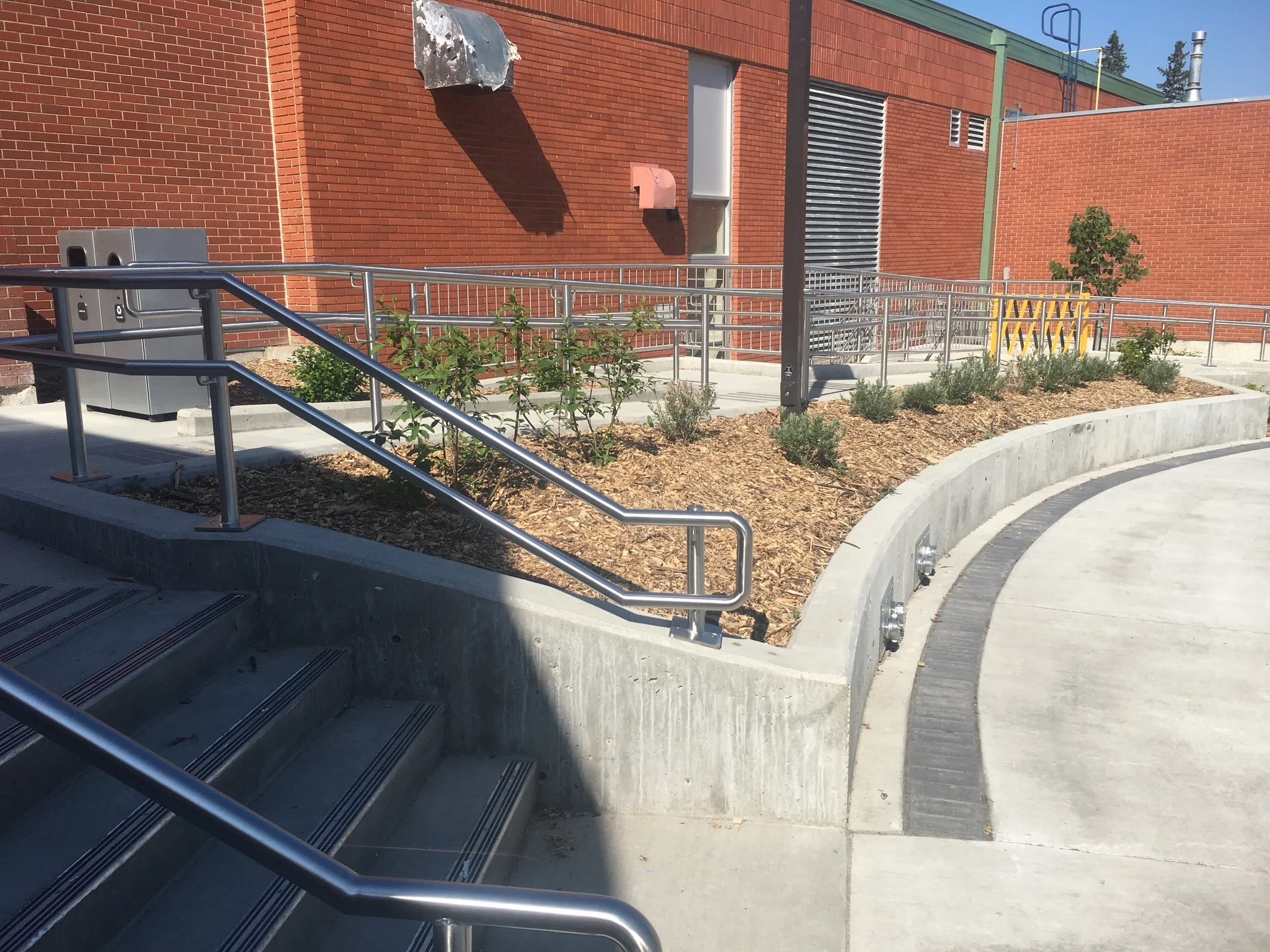 A concrete outdoor staircase and ramp with metal railings next to a landscaped area with small plants and mulch, adjacent to a red brick building with vents and pipes.