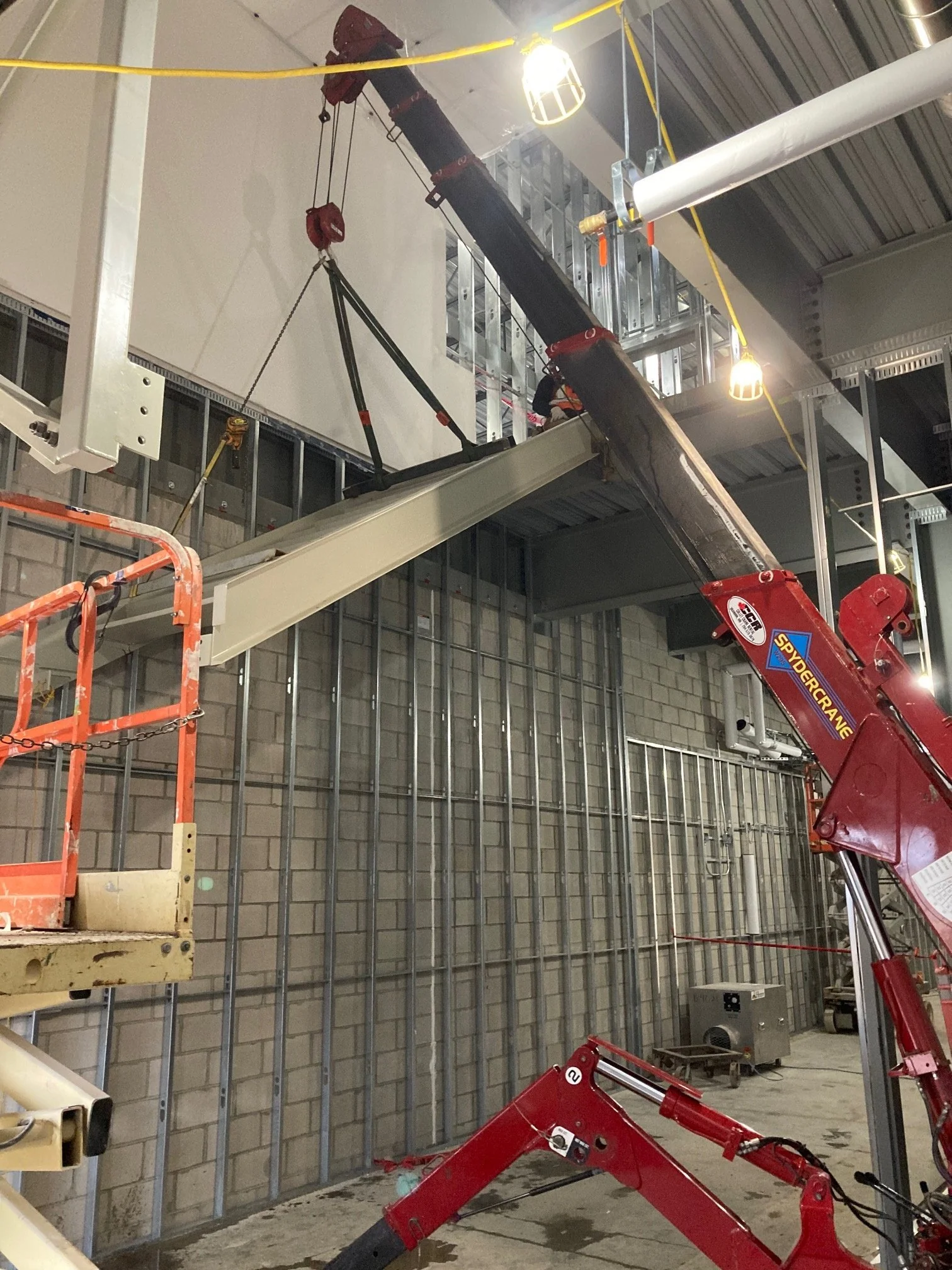 Construction site with a red crane lifting a large structural beam indoors, with scaffolding and metal framework nearby.