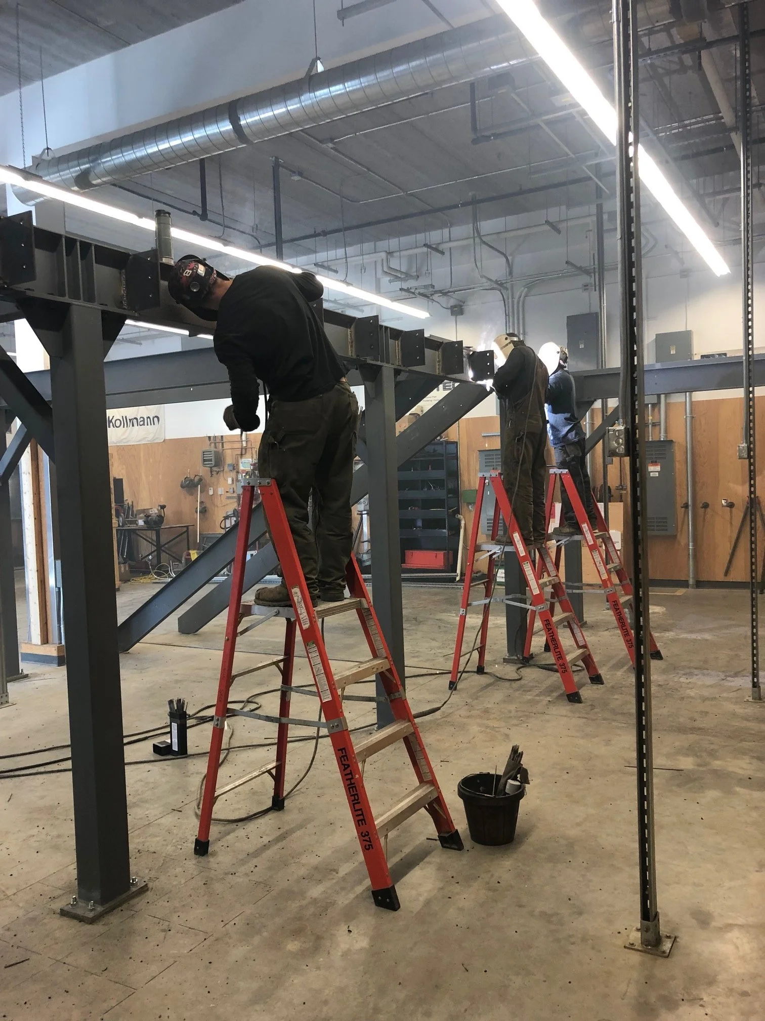Three workers on ladder scaffolding welding a large metal structure inside a workshop with ductwork and lighting overhead.