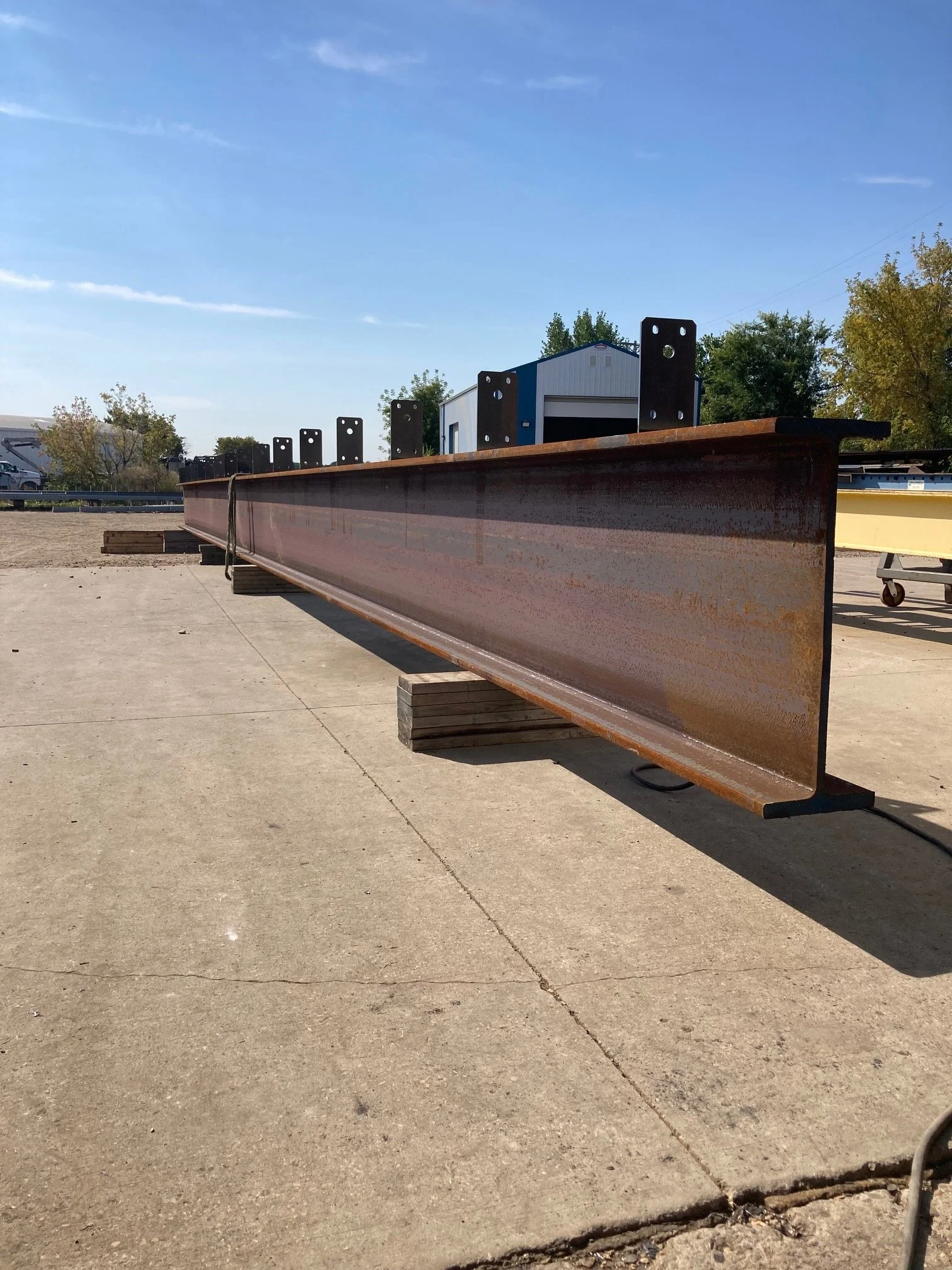 A large rusty steel I-beam supported by wooden blocks outdoors on a concrete surface, with trees, a building, and a blue sky in the background.