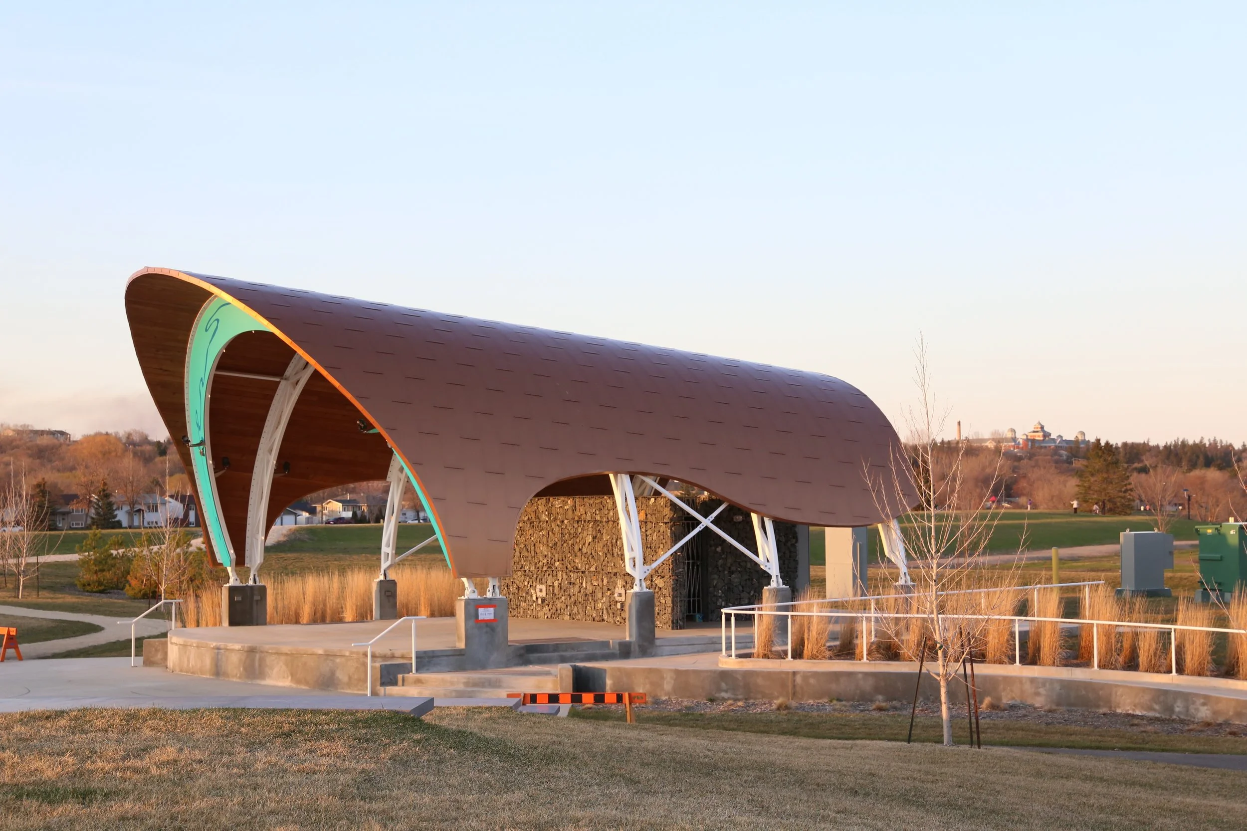 An outdoor stage with a curved wooden roof, supported by white metal beams, situated in a park during golden hour.