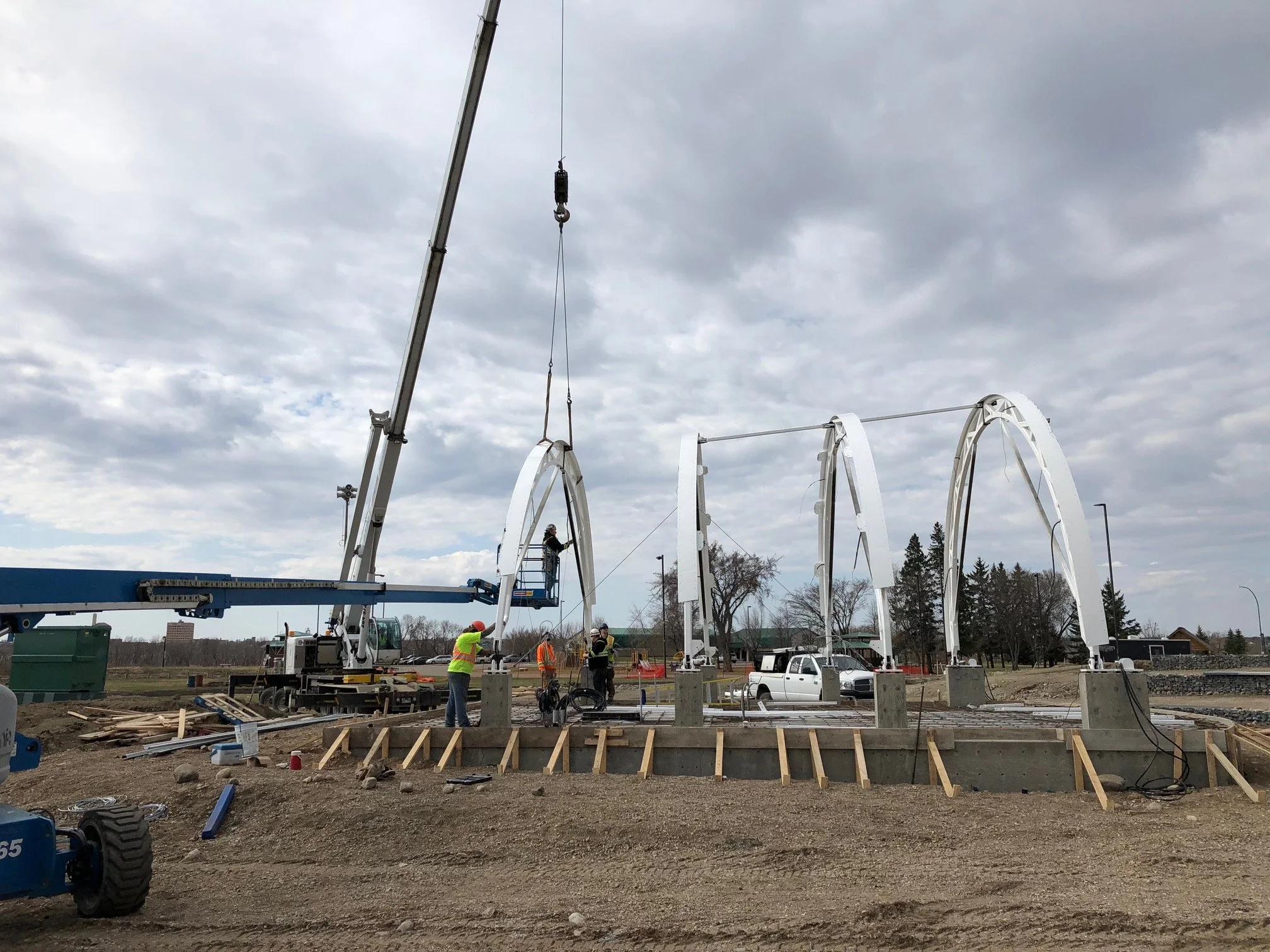 Construction site with large white arch structures being assembled, workers in safety vests and helmets, crane lifting parts, and a cloudy sky overhead.