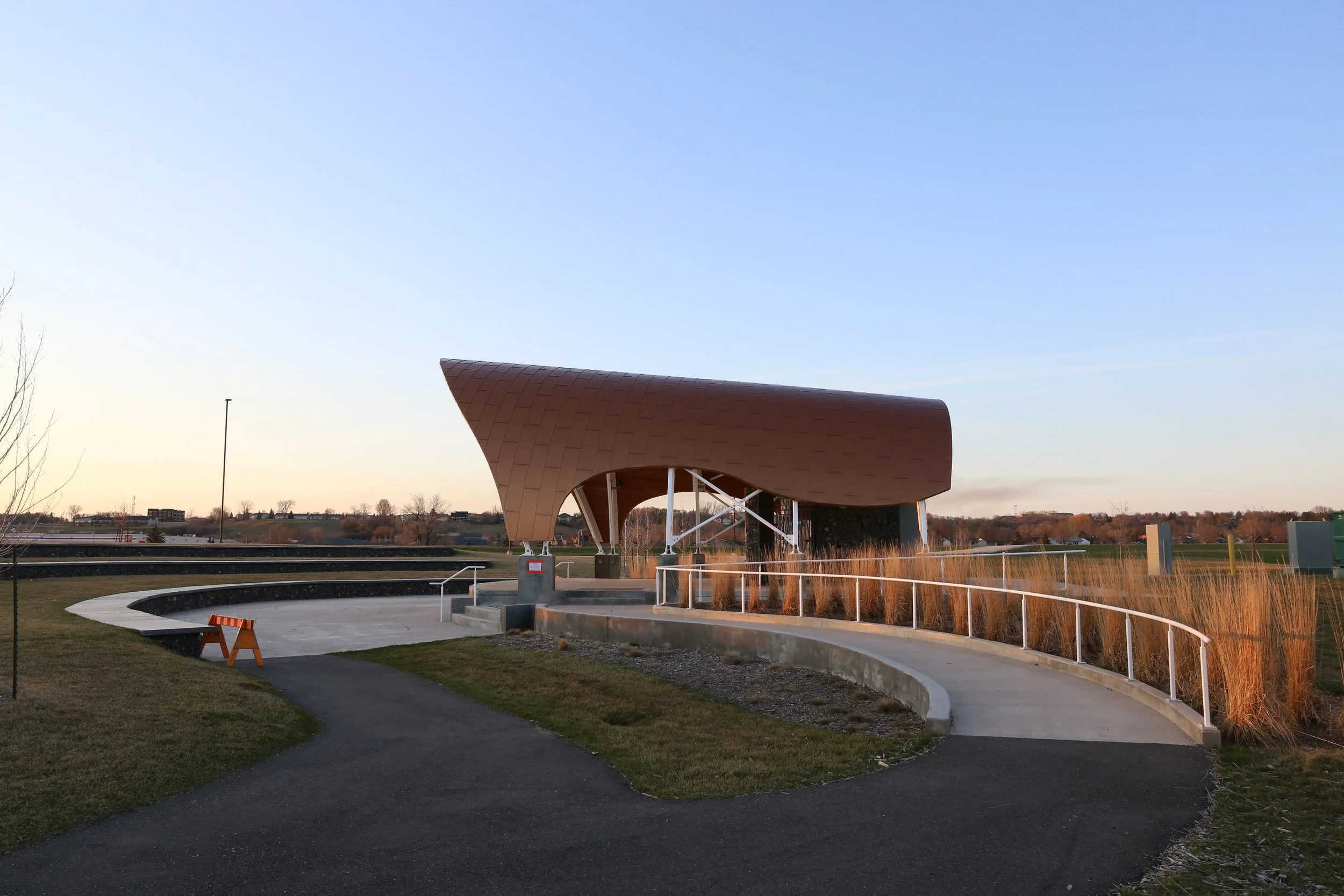 Modern architectural structure with a curved copper-colored roof, surrounded by a landscaped park with a paved walkway, grass, and tall ornamental grasses, under a clear evening sky.