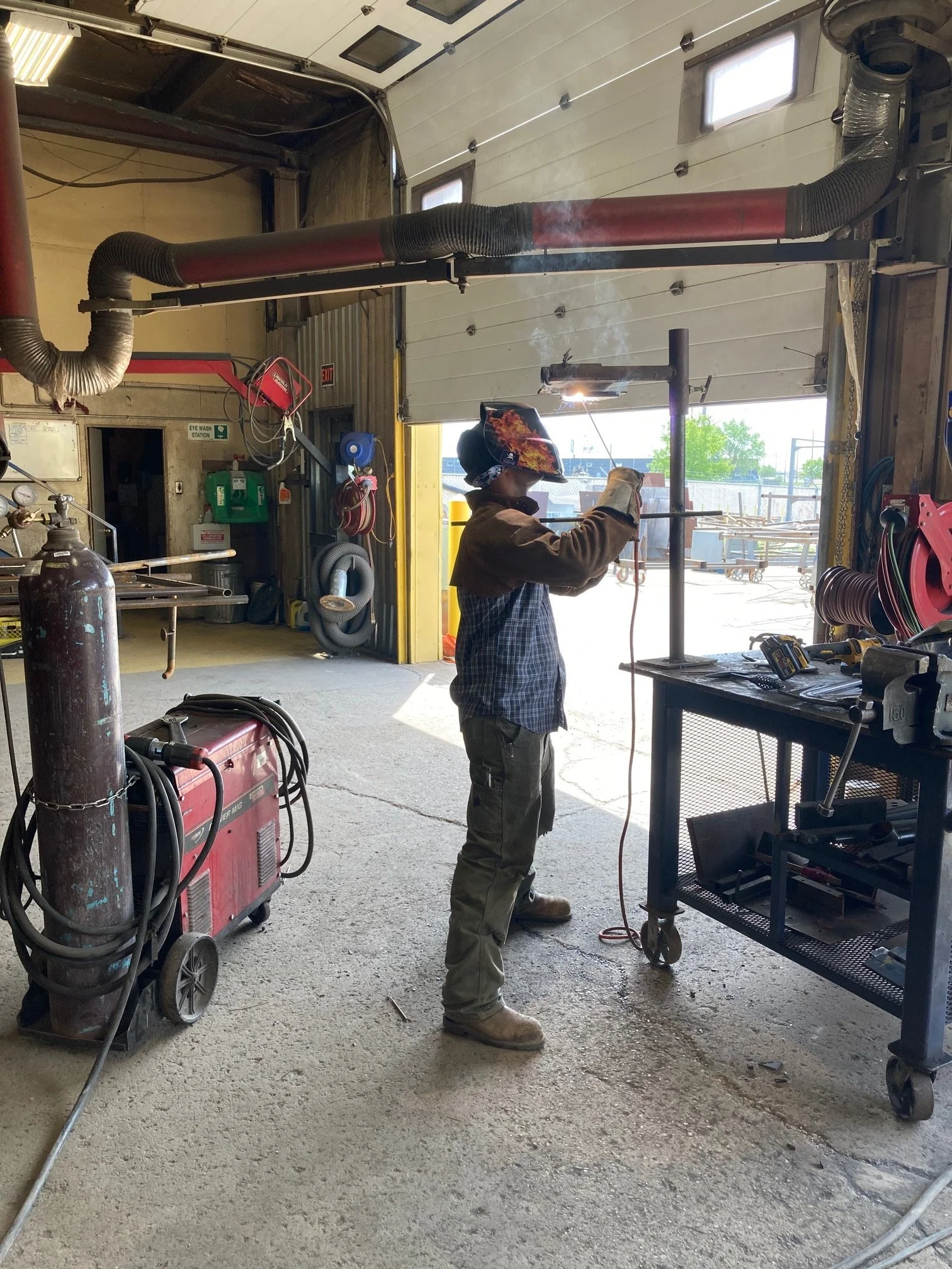 A worker welding metal pipe in a workshop with tools and equipment around, wearing protective gear including a welding helmet and gloves.