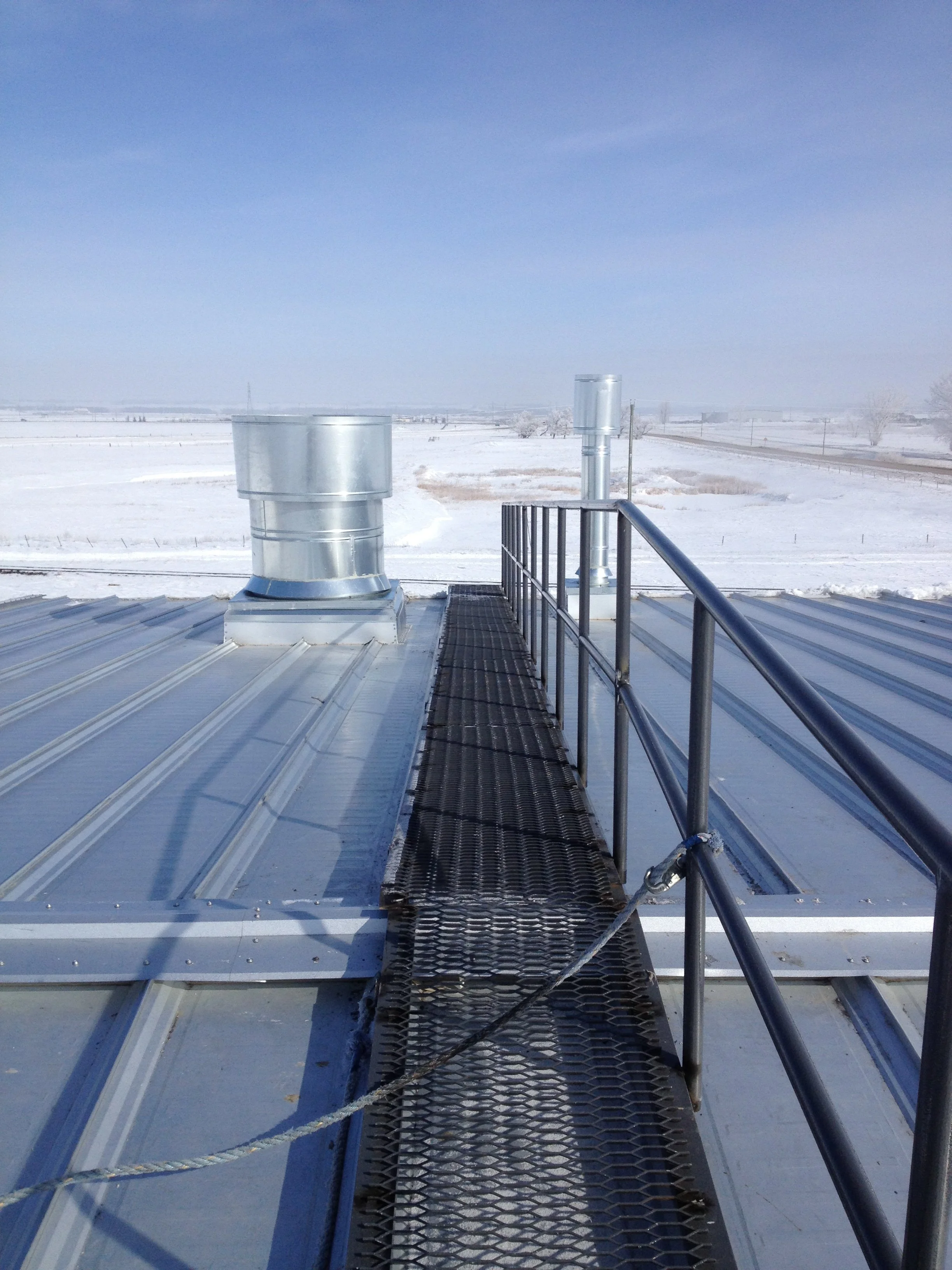 Metal roof with a black safety walkway and metal railing, overlooking snow-covered landscape with clear blue sky.