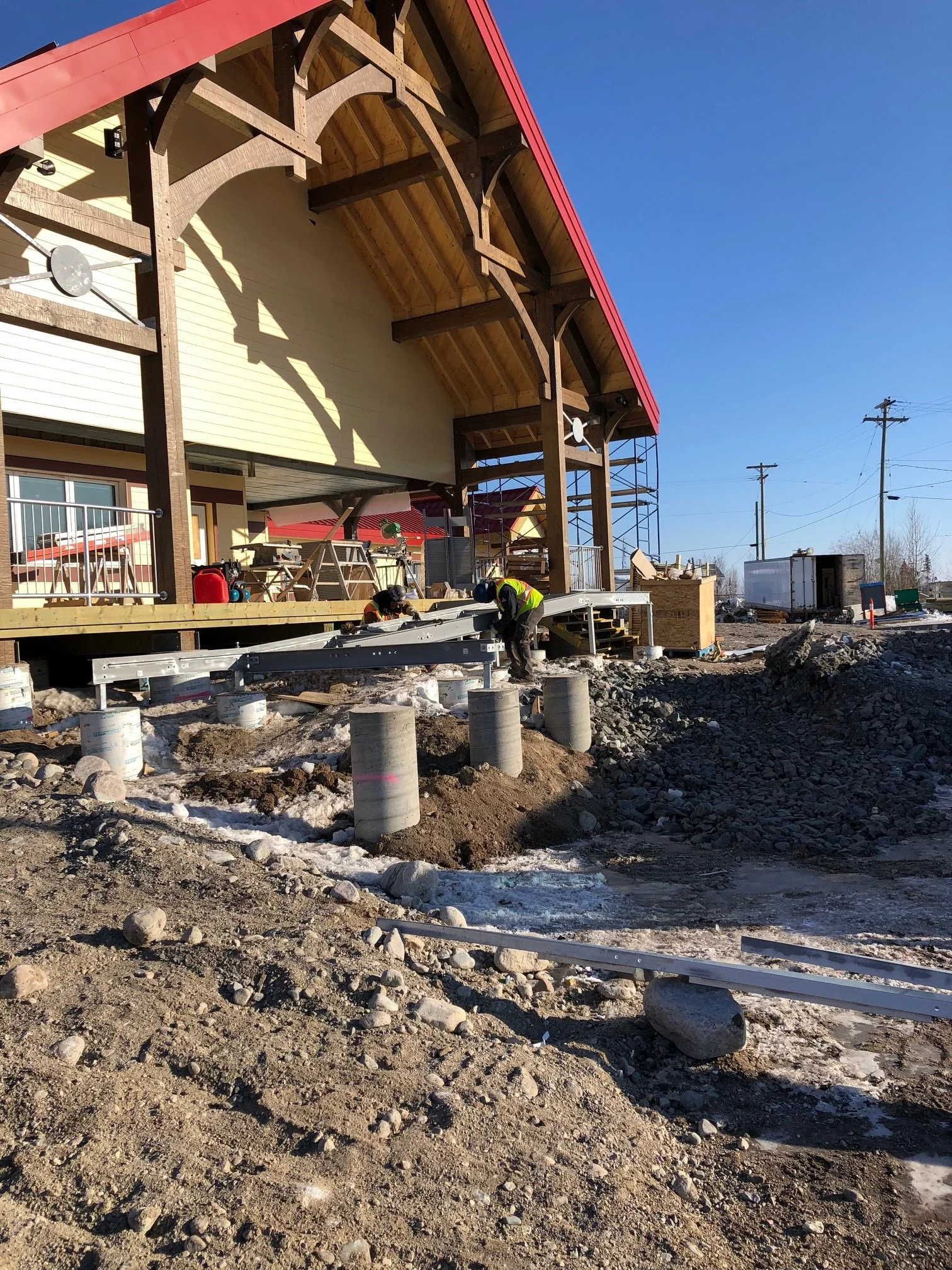 Construction workers install a steel beam in front of a large building with a wooden and metal framework, red roof trim, and scaffolding, with utility poles and trucks in the background.