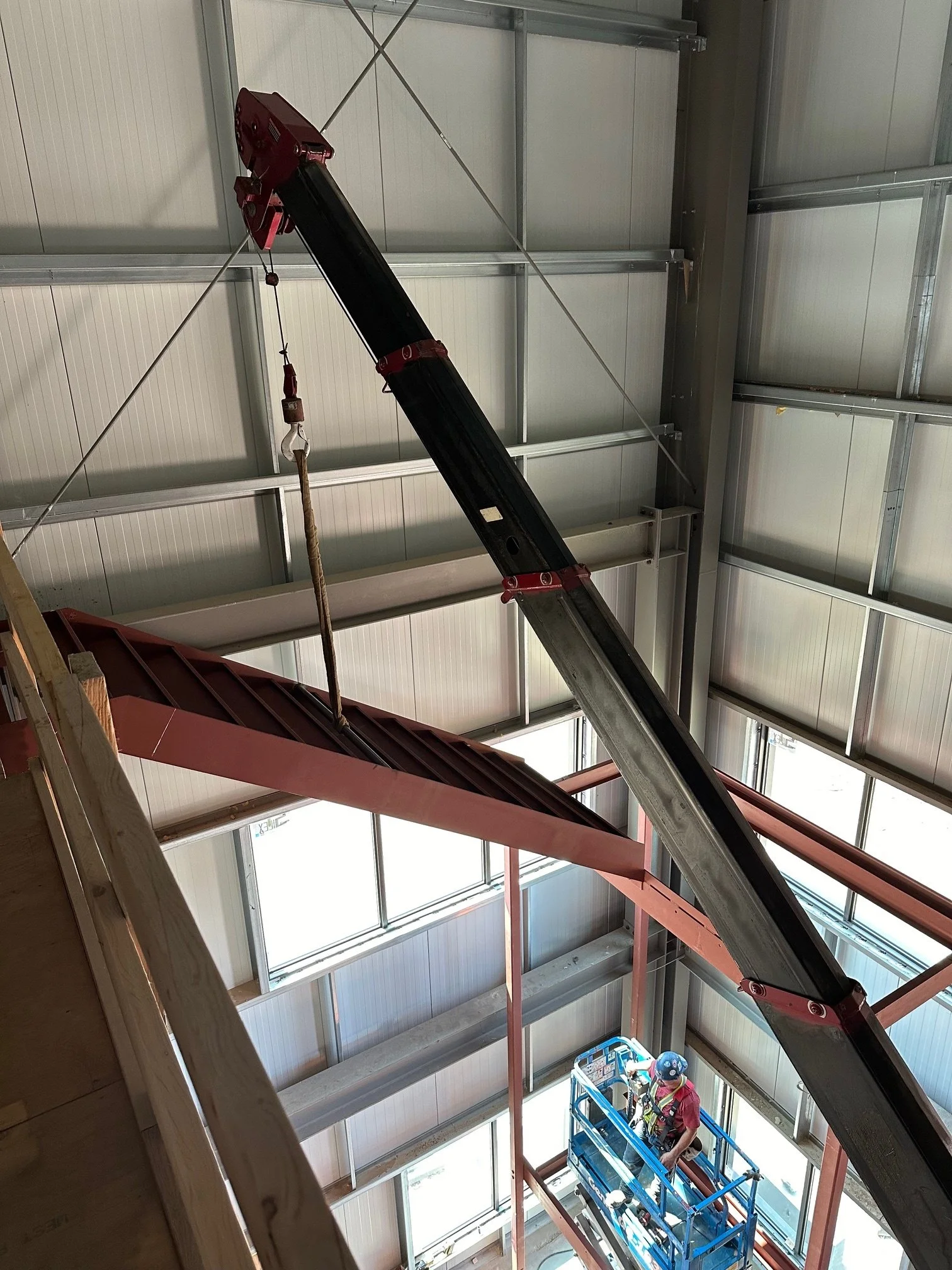 Construction worker in a safety harness and helmet operating a hydraulic lift inside a building under construction, with a crane visible overhead.