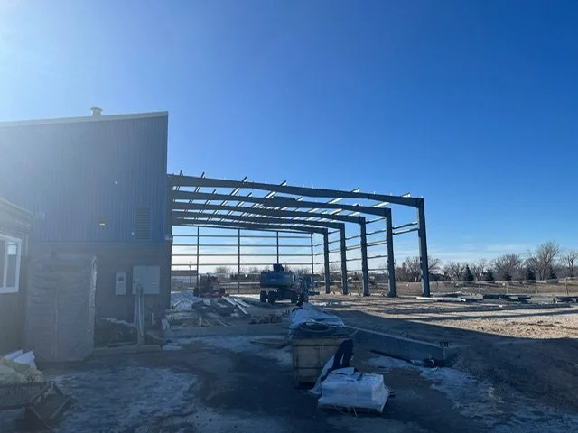 Construction site with steel framework for a building under clear blue sky, snow on the ground, and construction materials scattered around.
