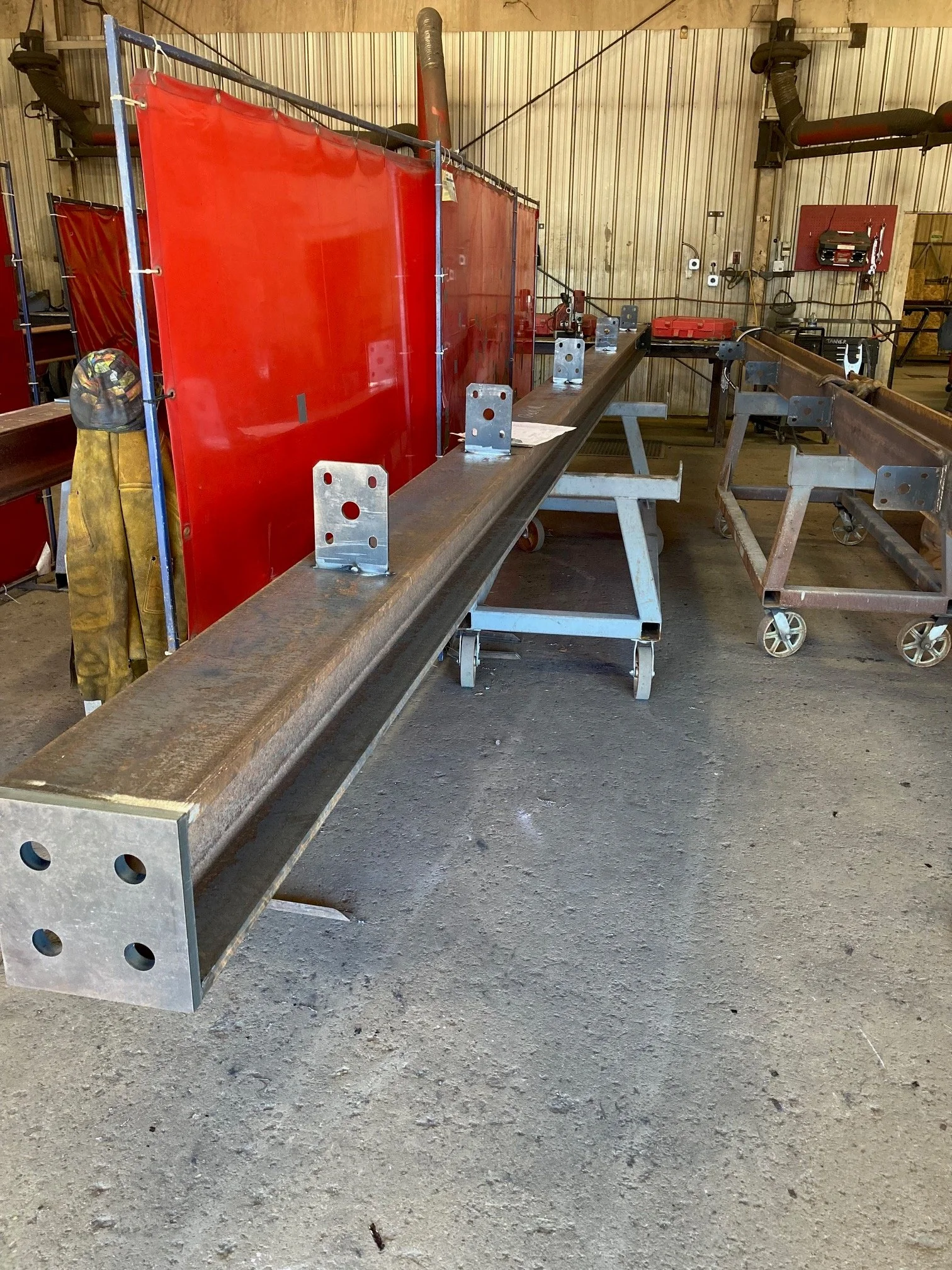 A workshop with a long metal beam supported by metal brackets on sawhorses, with welding separation screens, and tools in the background.