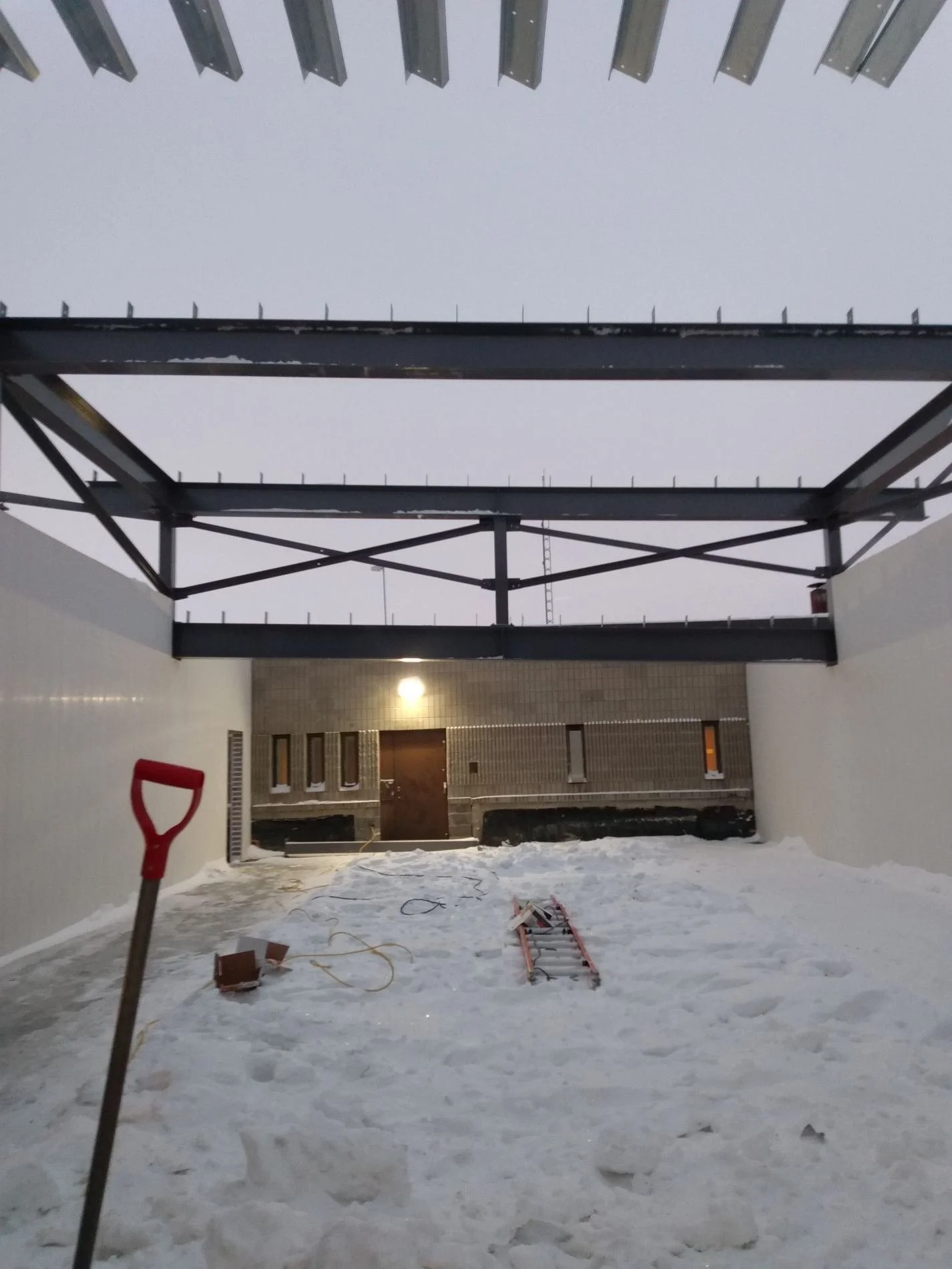Snow-covered rooftop with construction tools, ladder, shovel, and brick on the ground, with a building and a bright outdoor light in the background.