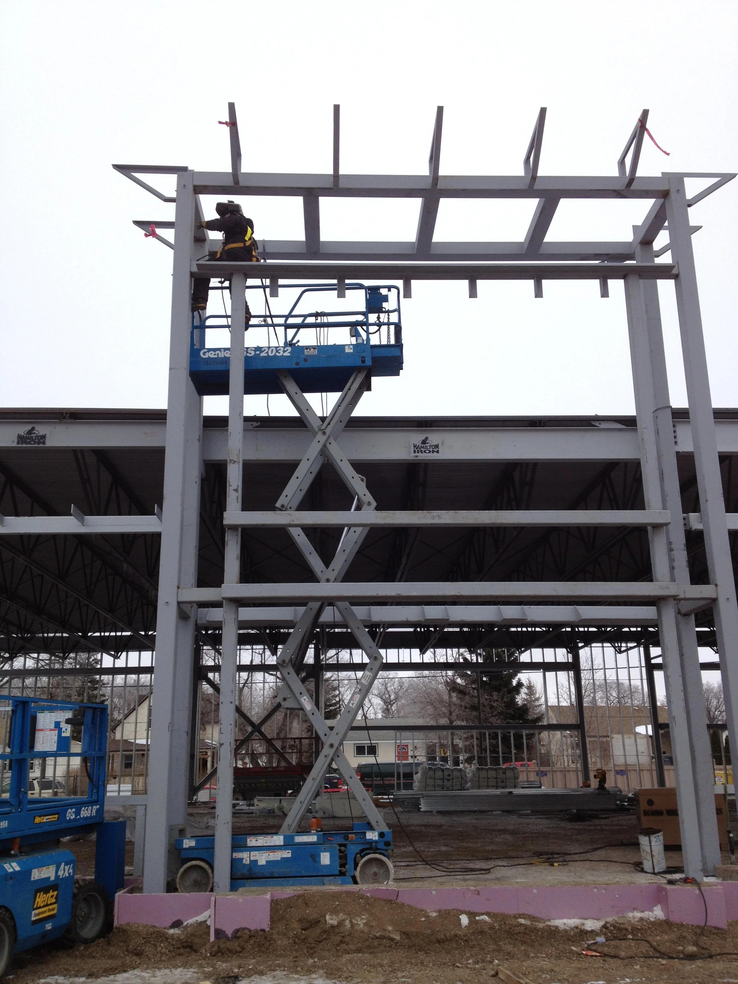 Construction worker on blue scissor lift assembling a metal framework in a warehouse or gymnasium with an outdoor background.