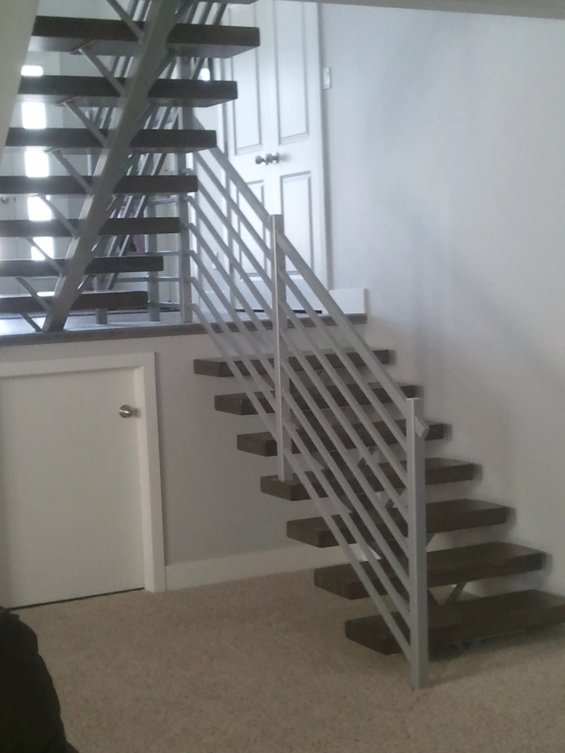 Interior view of a staircase with dark wooden steps and white metal railings, leading up to a second floor. There is a closed white door at the top of the stairs.