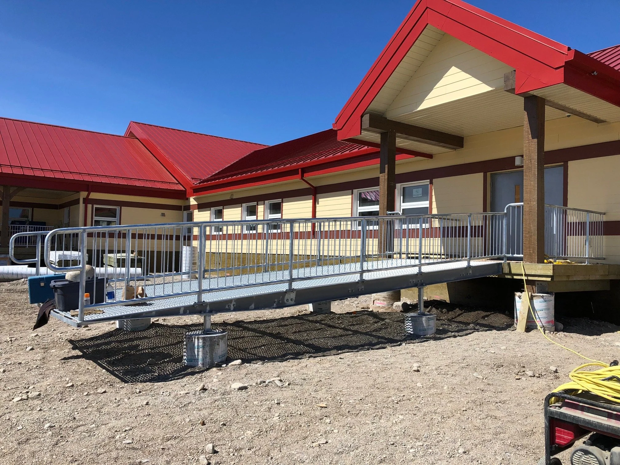 Construction site with a long metal wheelchair ramp in front of a yellow building with red roofing, supported by buckets and with construction tools nearby.
