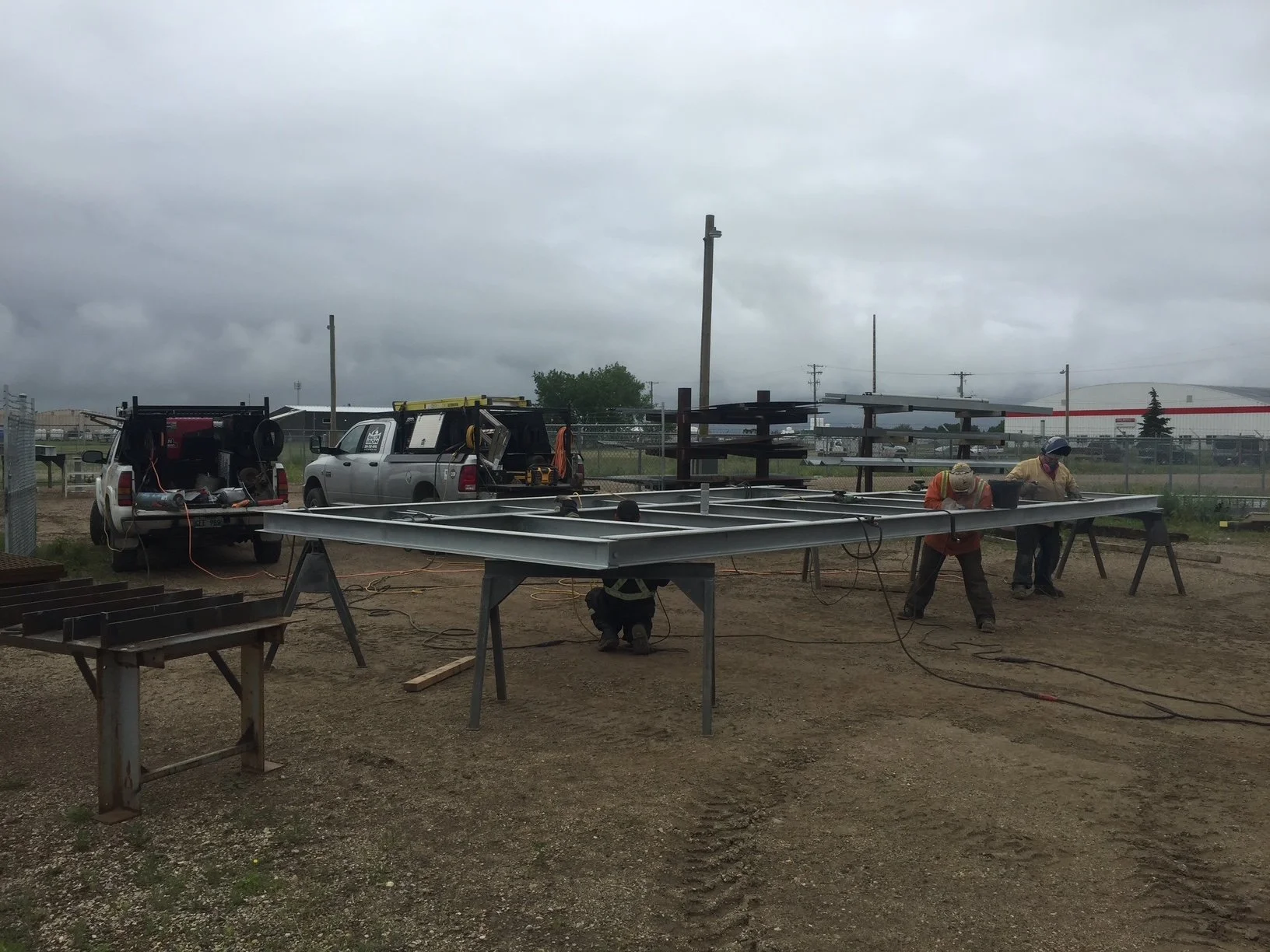 Construction workers assembling a metal structure outdoors on a cloudy day, with two pickup trucks and tools nearby.