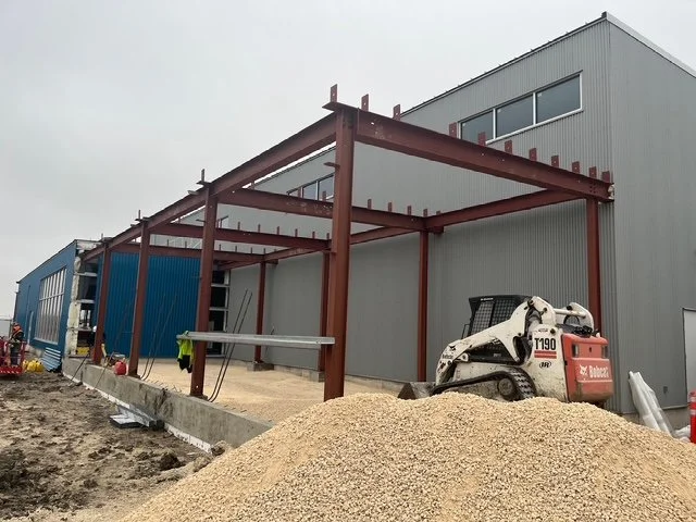 Construction site with steel frame structure being built adjacent to a gray building, a Bobcat skid-steer loader, and a large pile of gravel