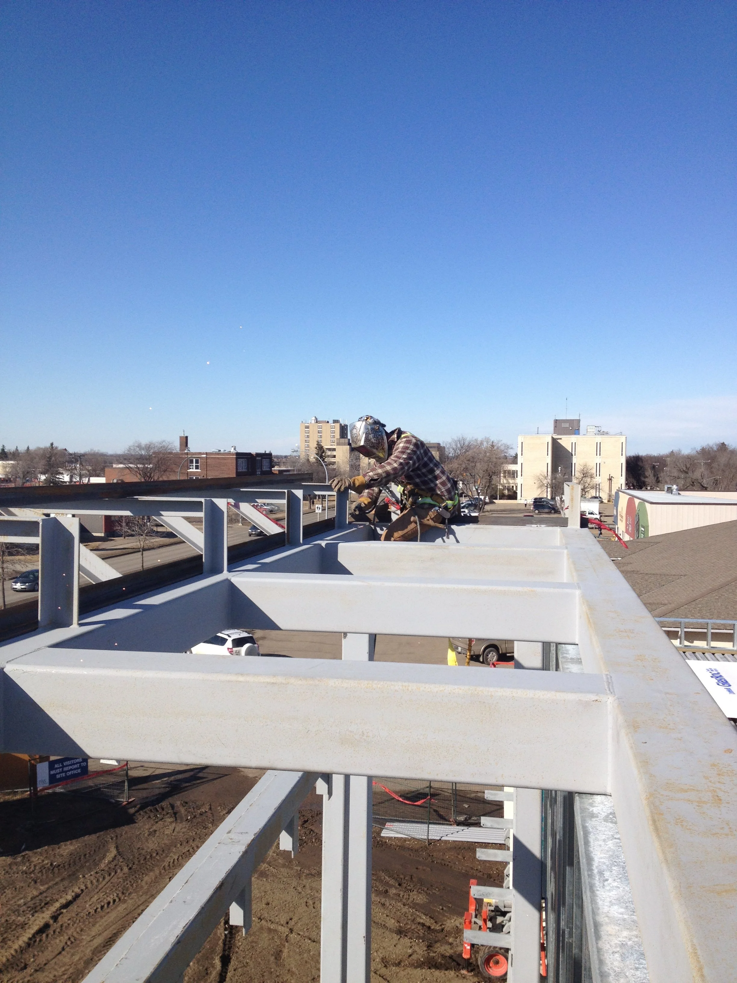 Worker wearing a safety helmet welding on a metal structure at the top of a building under construction with a clear blue sky in the background.