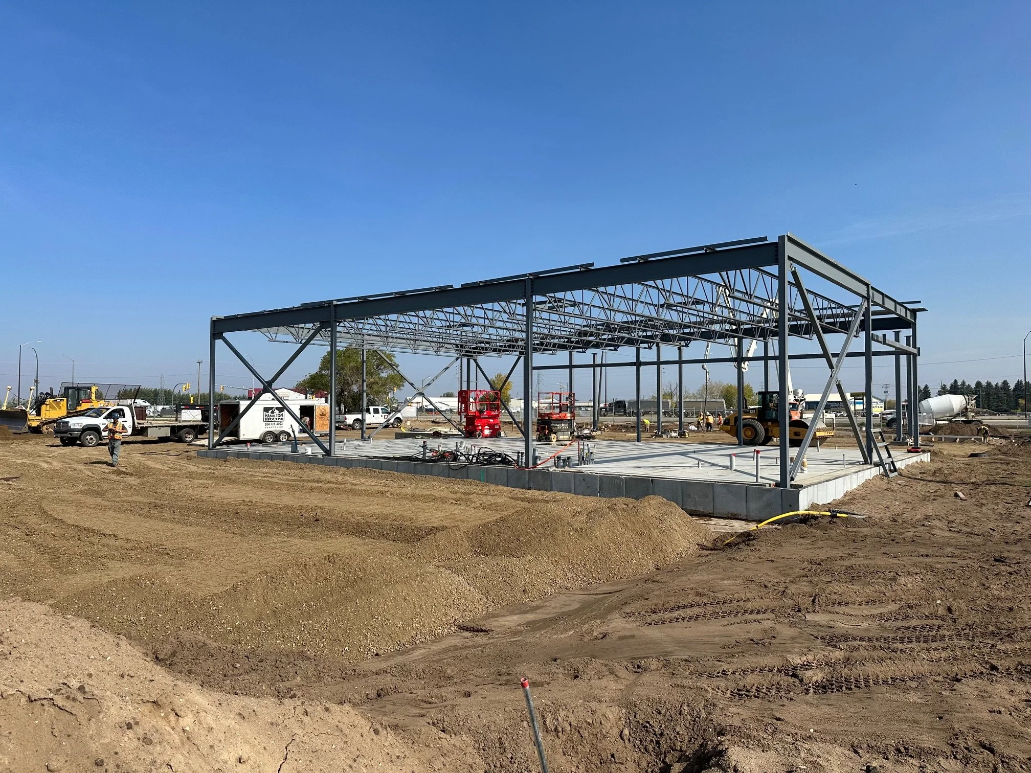 Steel framework for a building under construction on a dirt lot, with construction vehicles and workers in the background.