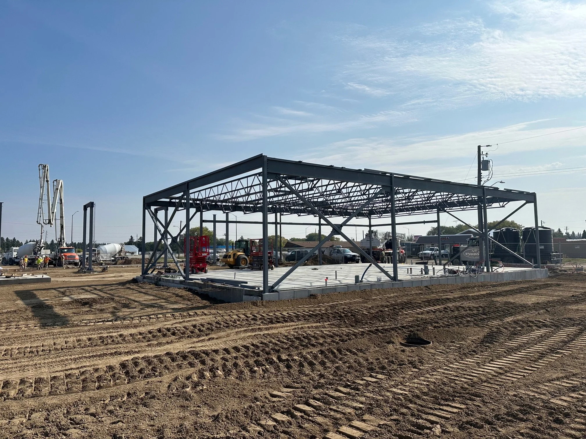 Construction site with a steel frame structure and construction equipment on a dirt lot during daytime.