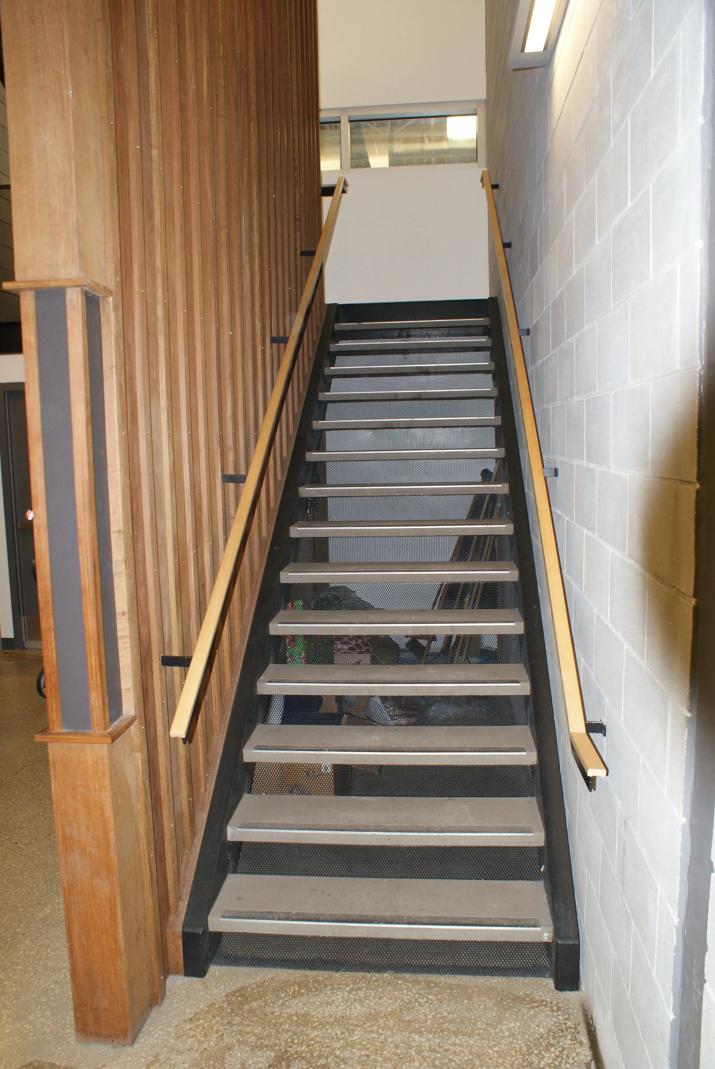 Indoor staircase with black metal frame, wooden handrails, and open risers, leading upwards in a building with wood paneling on one side and a white brick wall on the other.