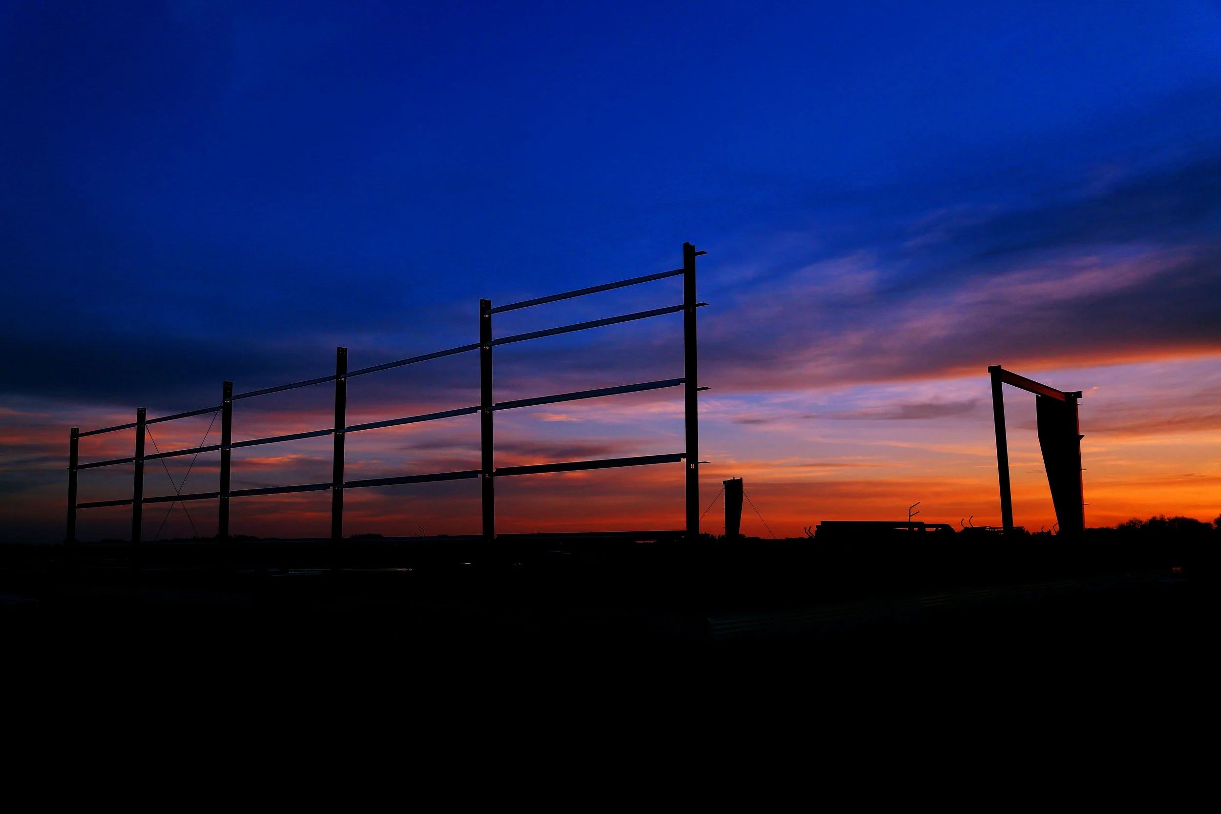 Silhouette of a construction site at sunset with empty framework and sky with shades of orange, pink, and blue.