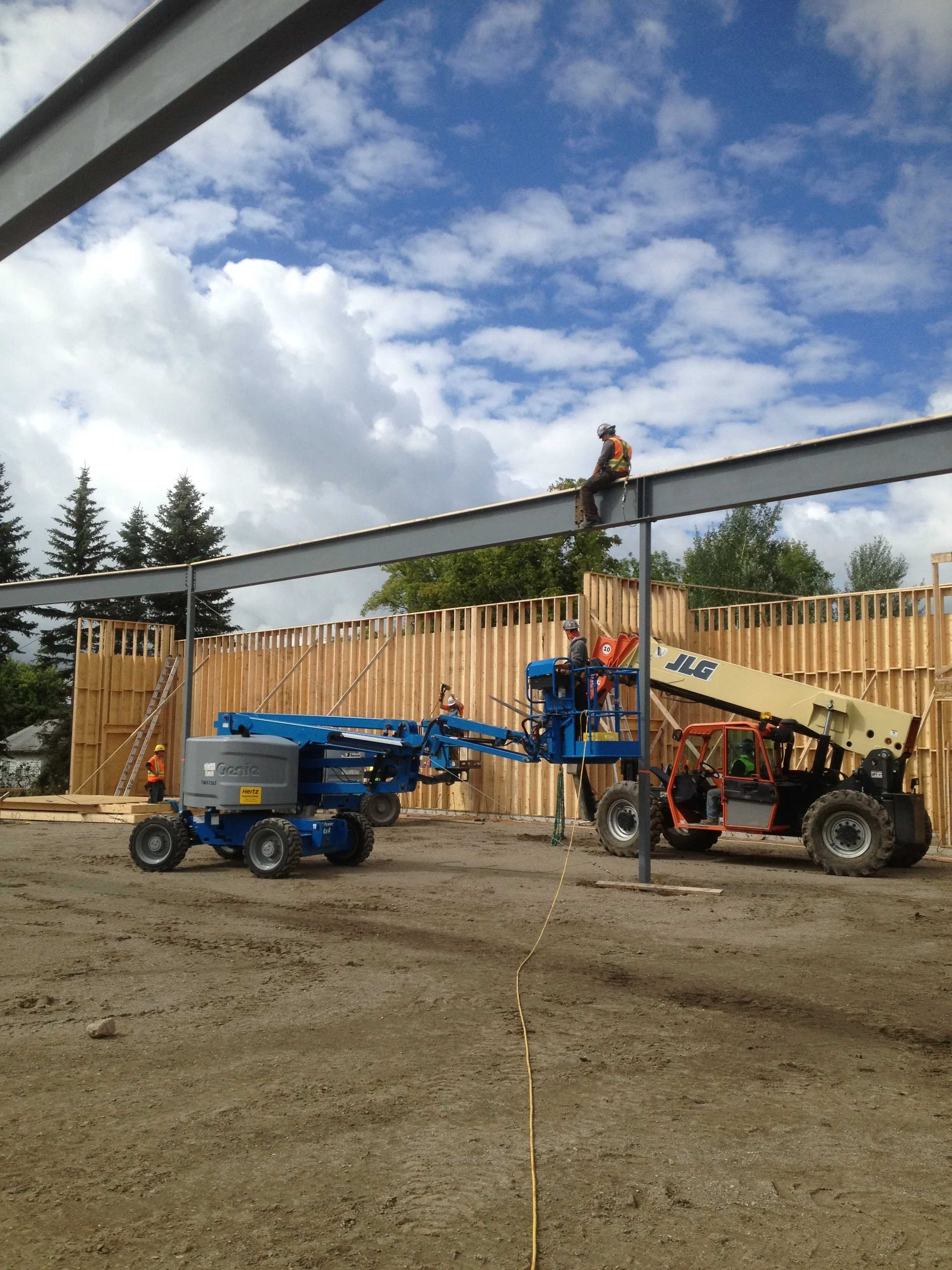 Construction workers building a wooden wall on a construction site with a metal beam structure overhead, surrounded by trees and a partly cloudy sky.