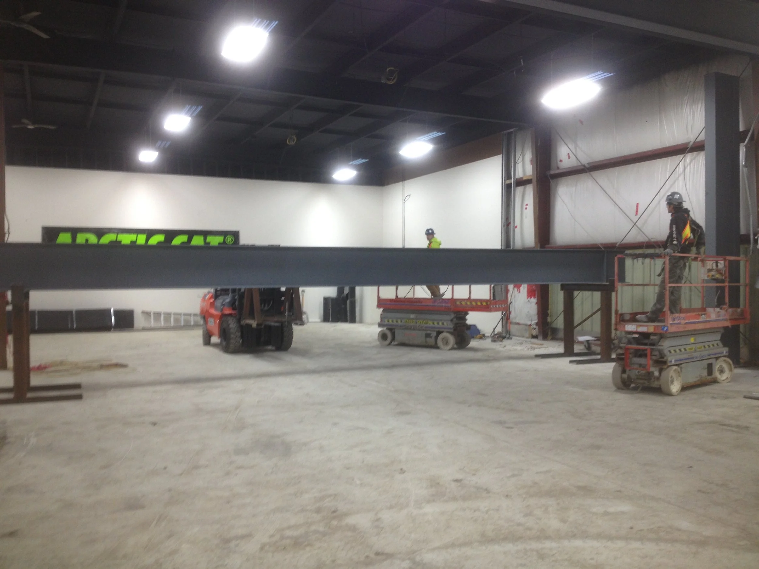 Interior view of a construction site with two workers on elevated platforms, a forklift, and a large steel beam being installed. The wall features a green sign reading 'ARCTIC CAT'.
