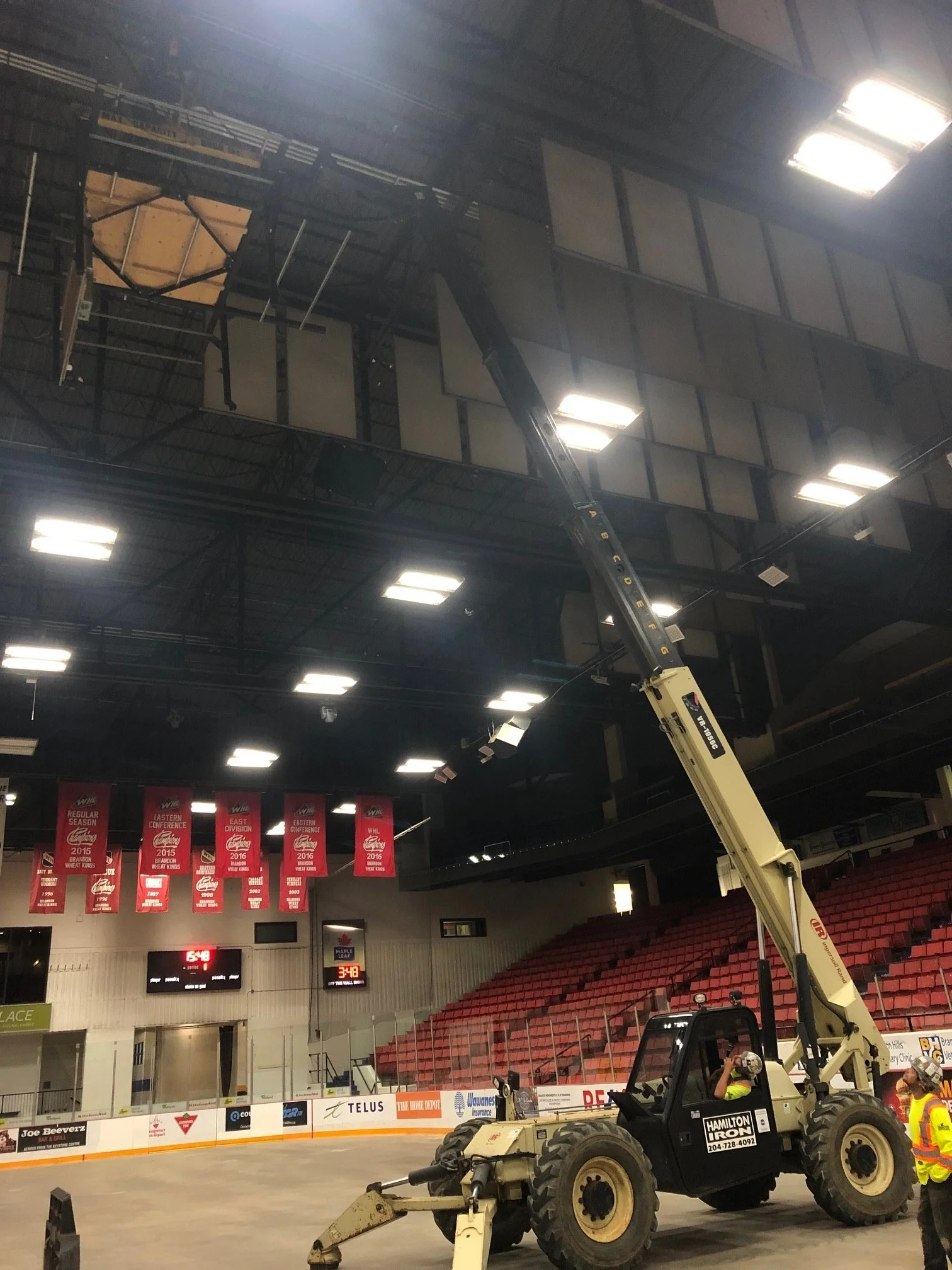 Construction vehicle inside a sports arena with red seats and banners hanging on the wall.