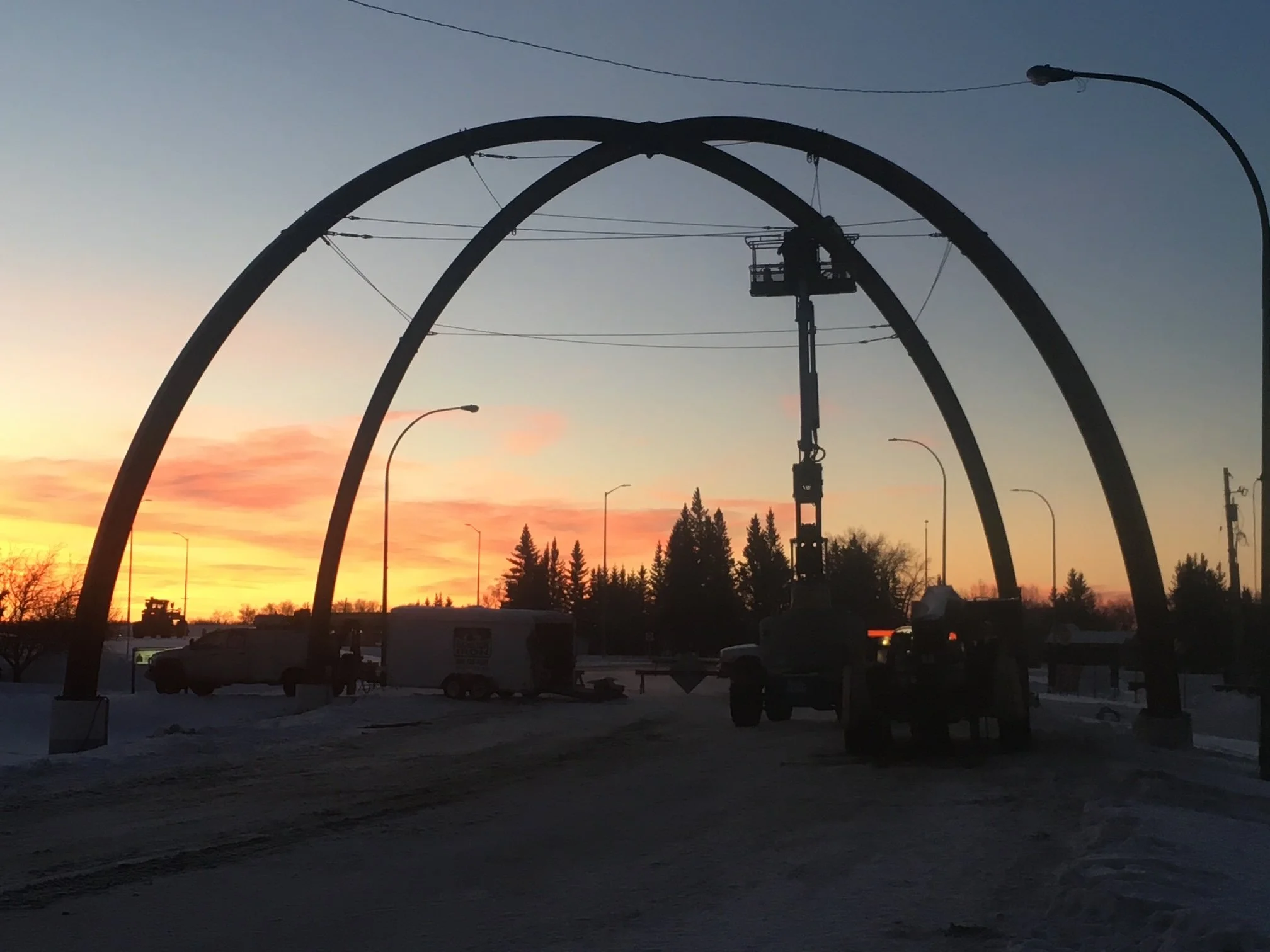 Construction workers installing large arches for a new structure against a sunset sky with colorful clouds and silhouetted trees.