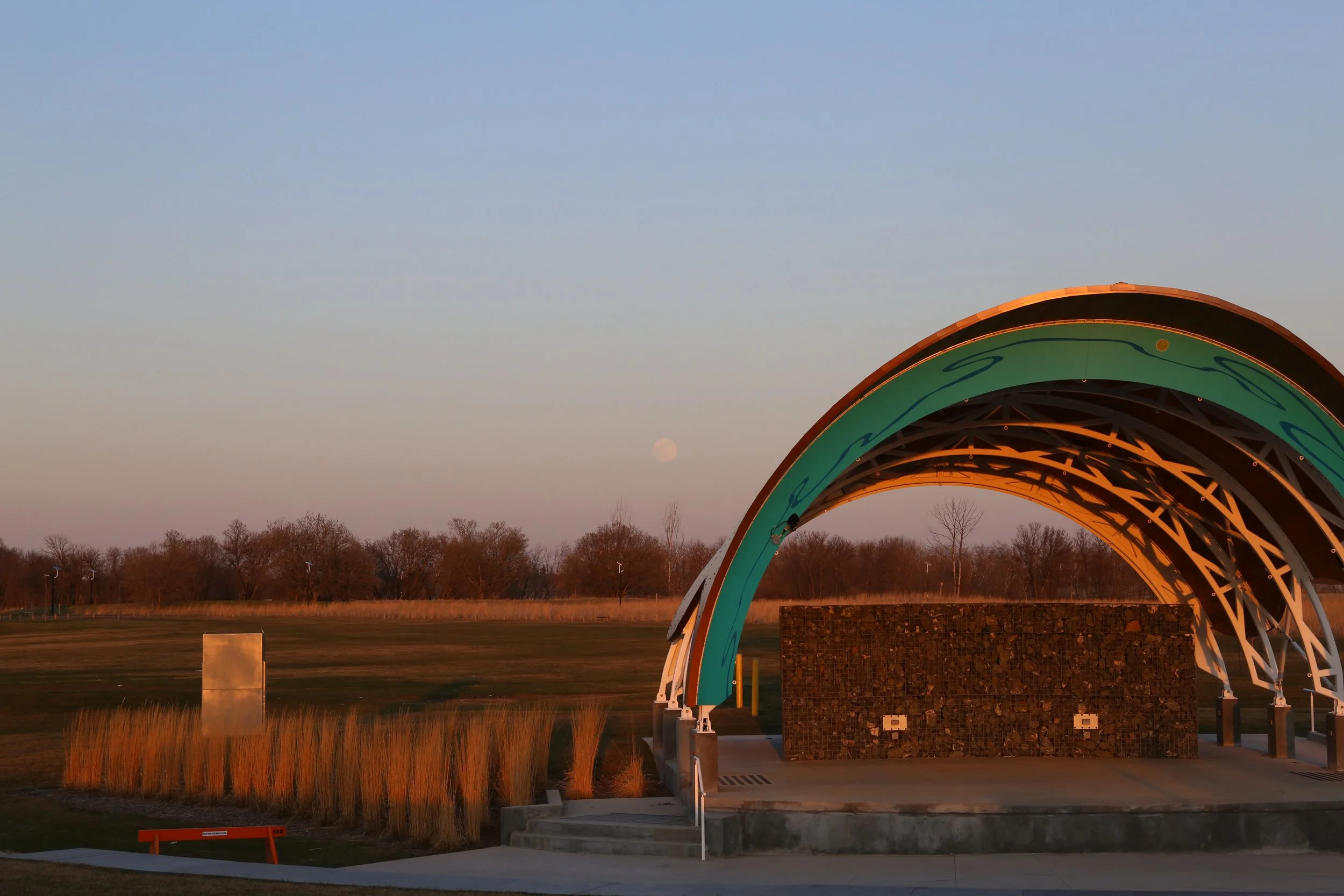 A modern outdoor amphitheater with a curved, colorful metal canopy structure, set in a park during sunset with a visible moon in the sky.